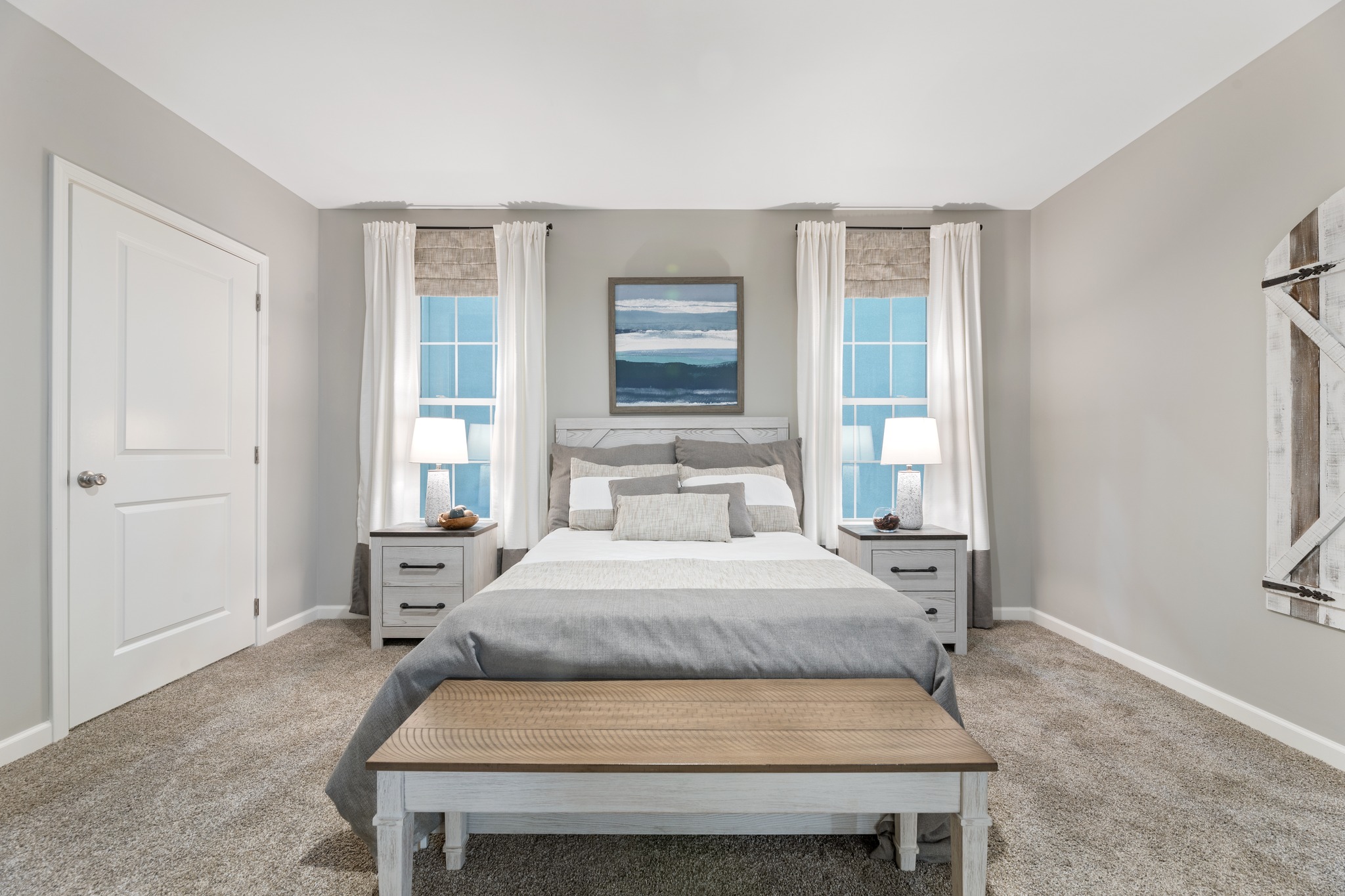 Cozy bedroom with a central bed, gray and white bedding, flanked by lamps on nightstands. Artwork above the bed, soft carpet, and a rustic bench at the foot.
