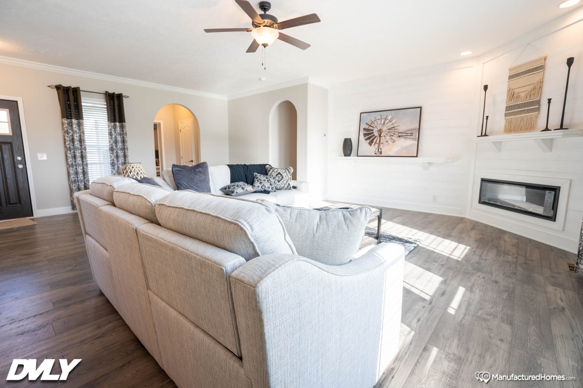 Spacious living room with a large beige sectional sofa, patterned cushions, and wooden floor. Ceiling fan, modern fireplace, and geometric art on walls. Cozy and inviting.