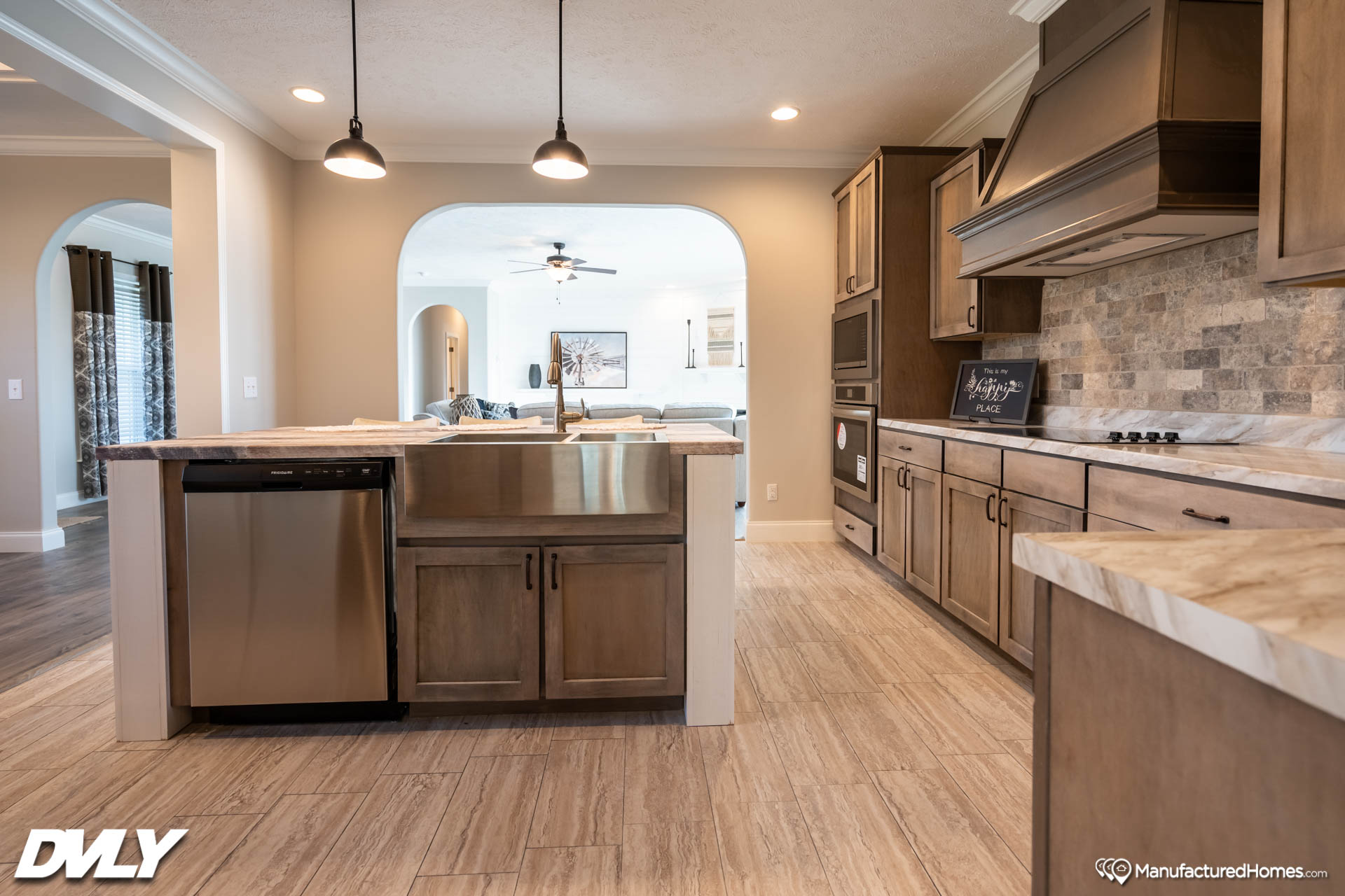Spacious kitchen with wooden cabinets, stone backsplash, and a large farmhouse sink. Bright, with pendant lights and an open view to the living room. Cozy tone.