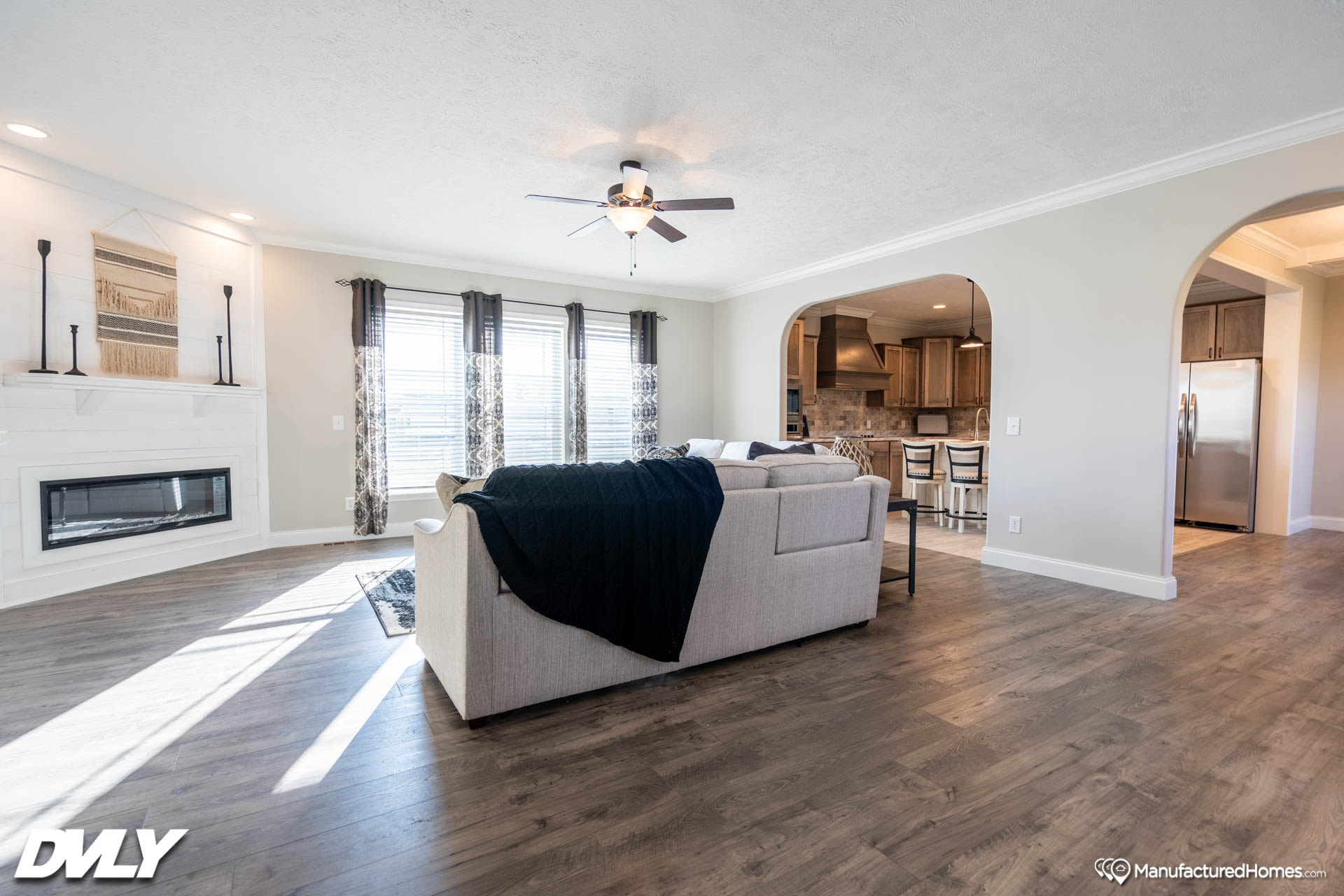 Spacious living room with wooden floor, beige sectional sofa, black throw blanket, large windows, ceiling fan, modern fireplace, and open arch to kitchen.