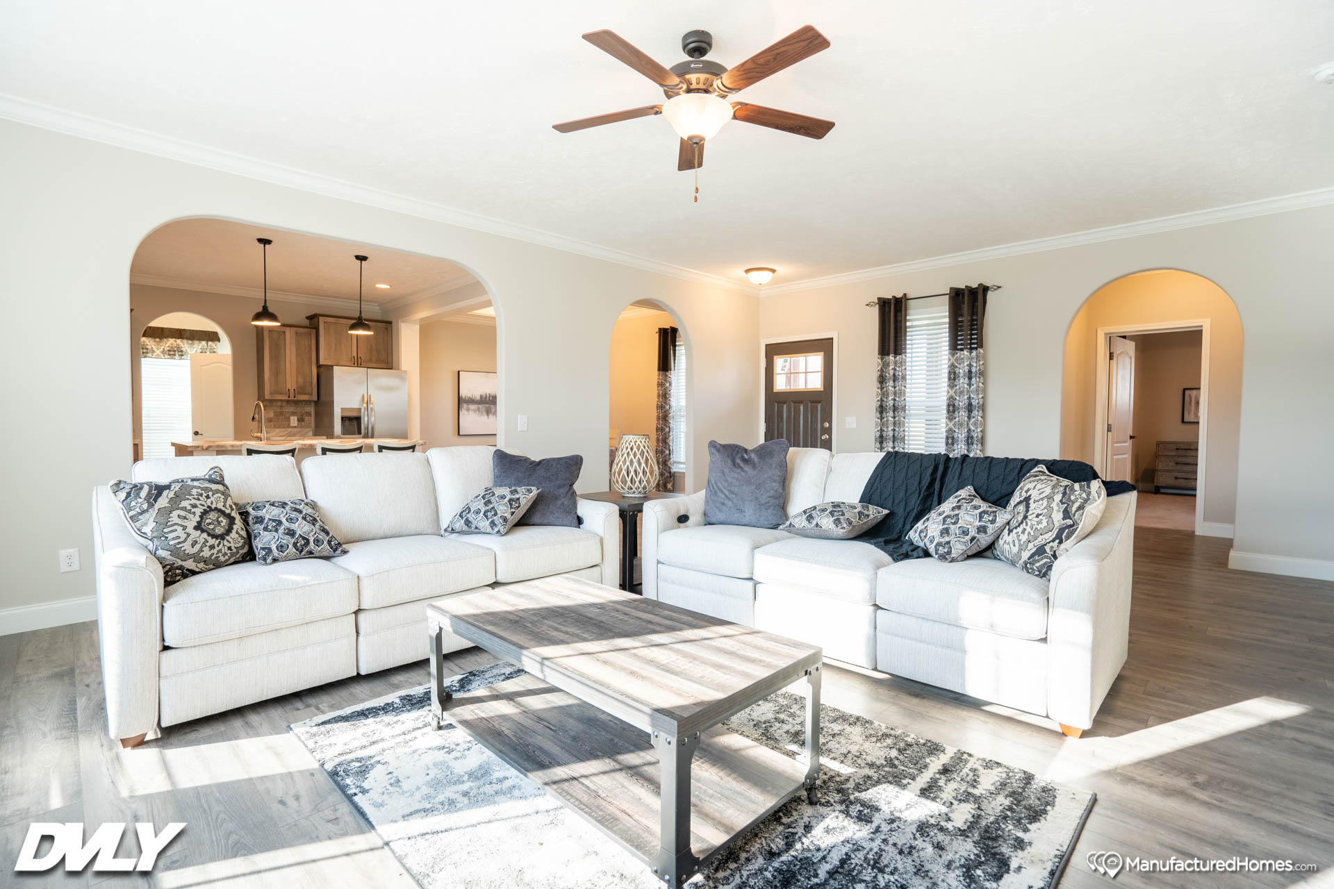 Bright living room with two white sofas adorned with patterned pillows. A wooden coffee table sits on a gray rug. Ceiling fan above and entryway visible. Cozy and inviting.