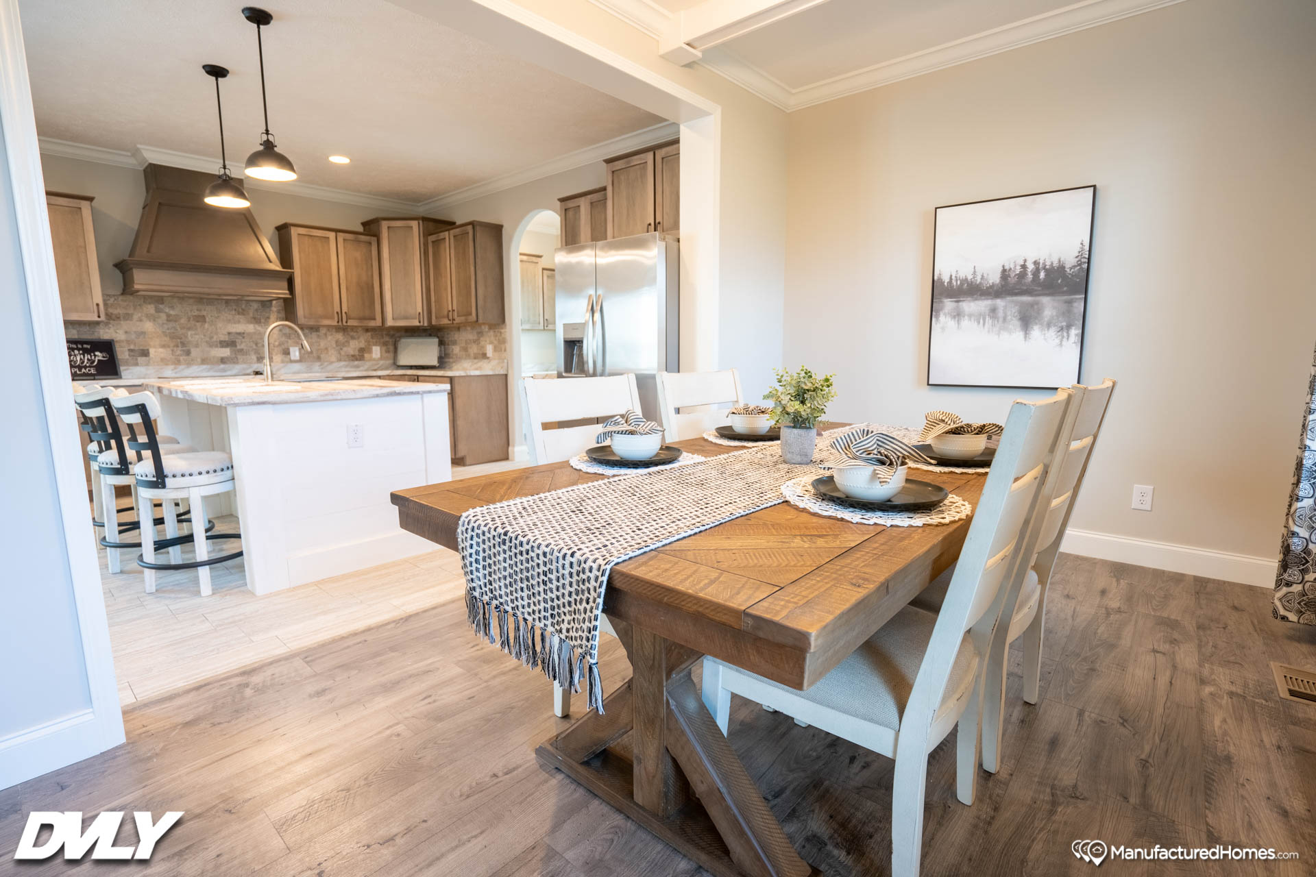 A cozy dining area with a wooden table, set for four, featuring white chairs and a patterned runner. Nearby is a modern kitchen with light wood cabinets.