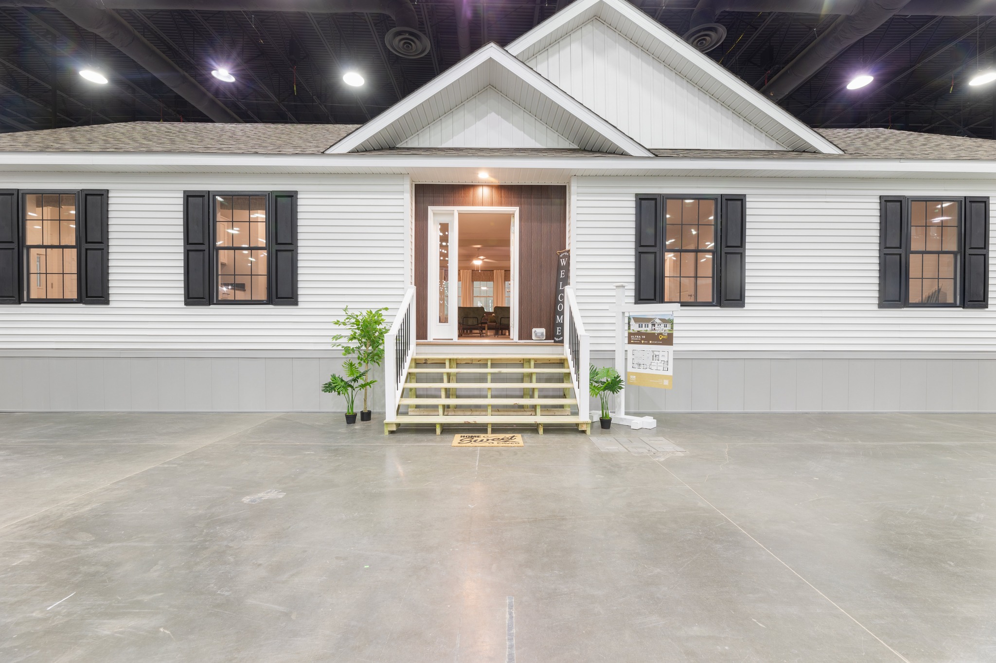 A white modular home with a gabled roof is displayed indoors. It has two front windows with black shutters, central wooden steps, and potted plants.
