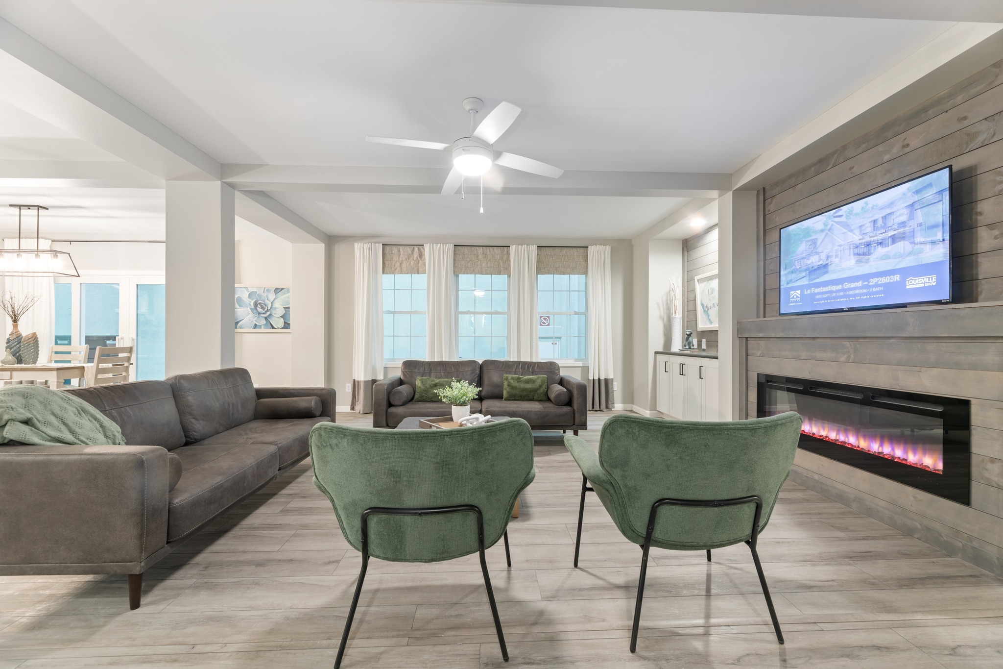 Contemporary living room with ceiling fan, gray sofas, green armchairs, wall-mounted TV, and electric fireplace. Bright and inviting ambiance.