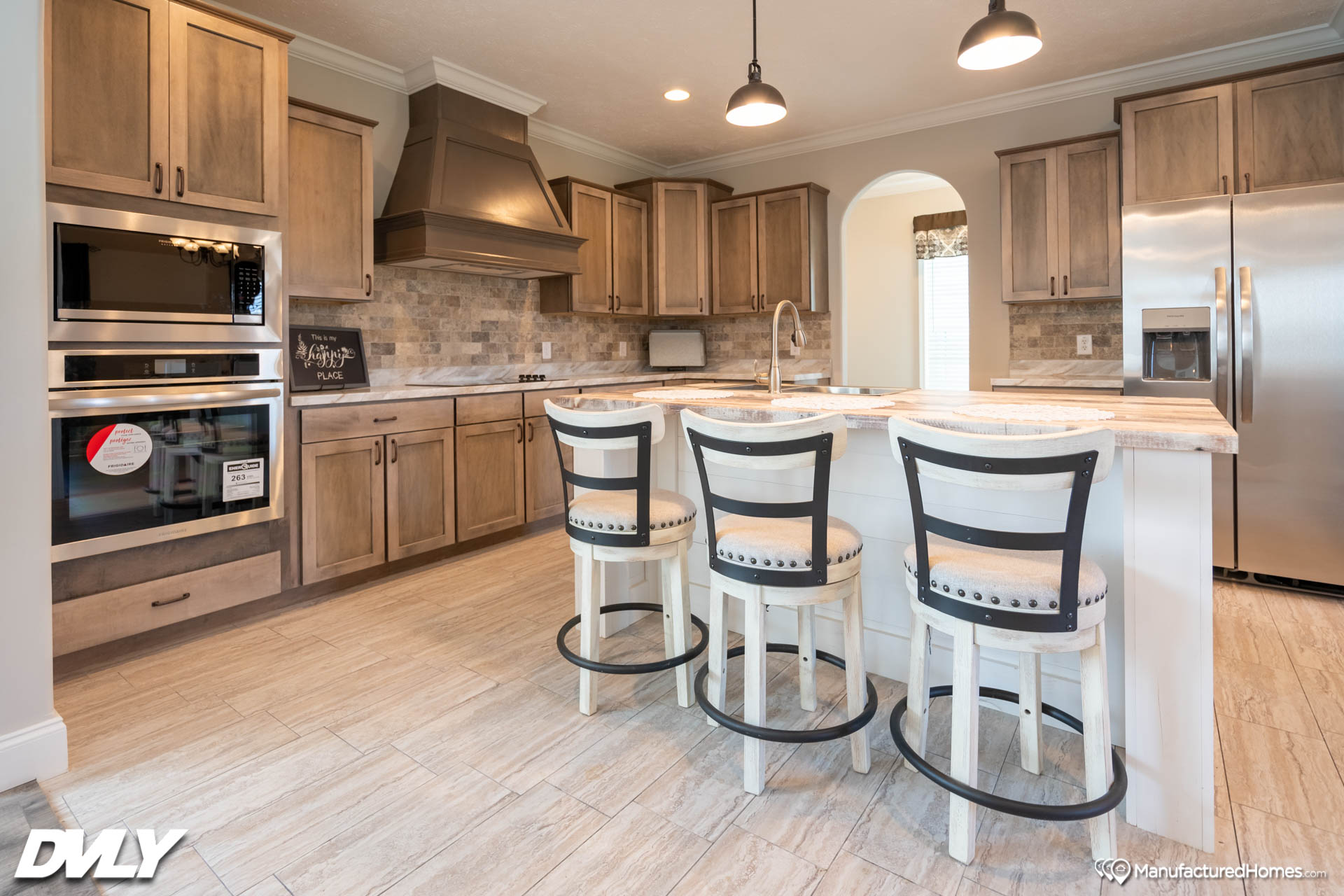 Modern kitchen with wooden cabinets, stainless steel appliances, and an island with three bar stools. Warm lighting and neutral tones create a cozy atmosphere.
