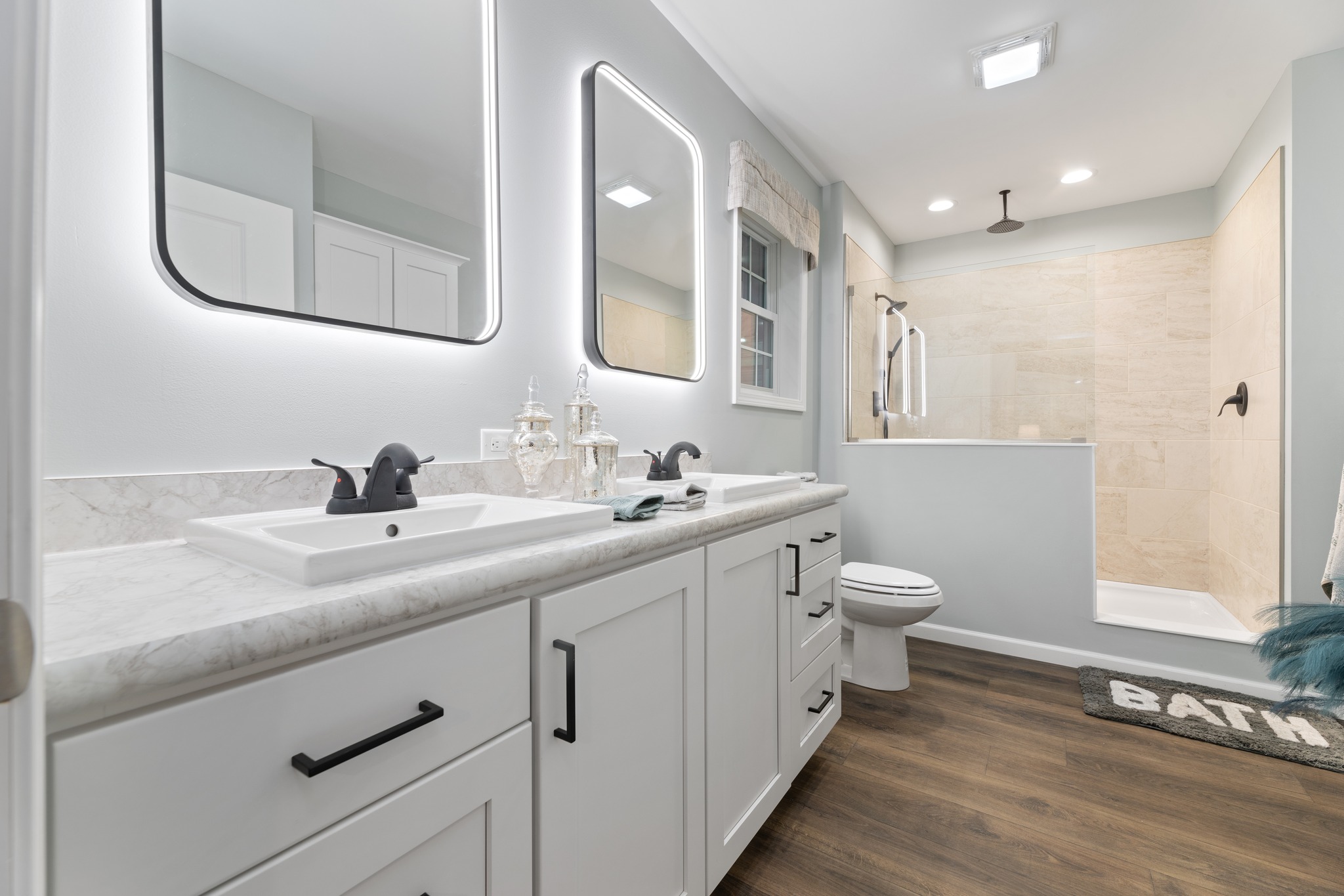A modern bathroom with a double vanity and illuminated mirrors, light marble countertop, and dark faucets. It has a walk-in shower, beige tiles, and wood flooring. A bath mat adds a cozy touch.