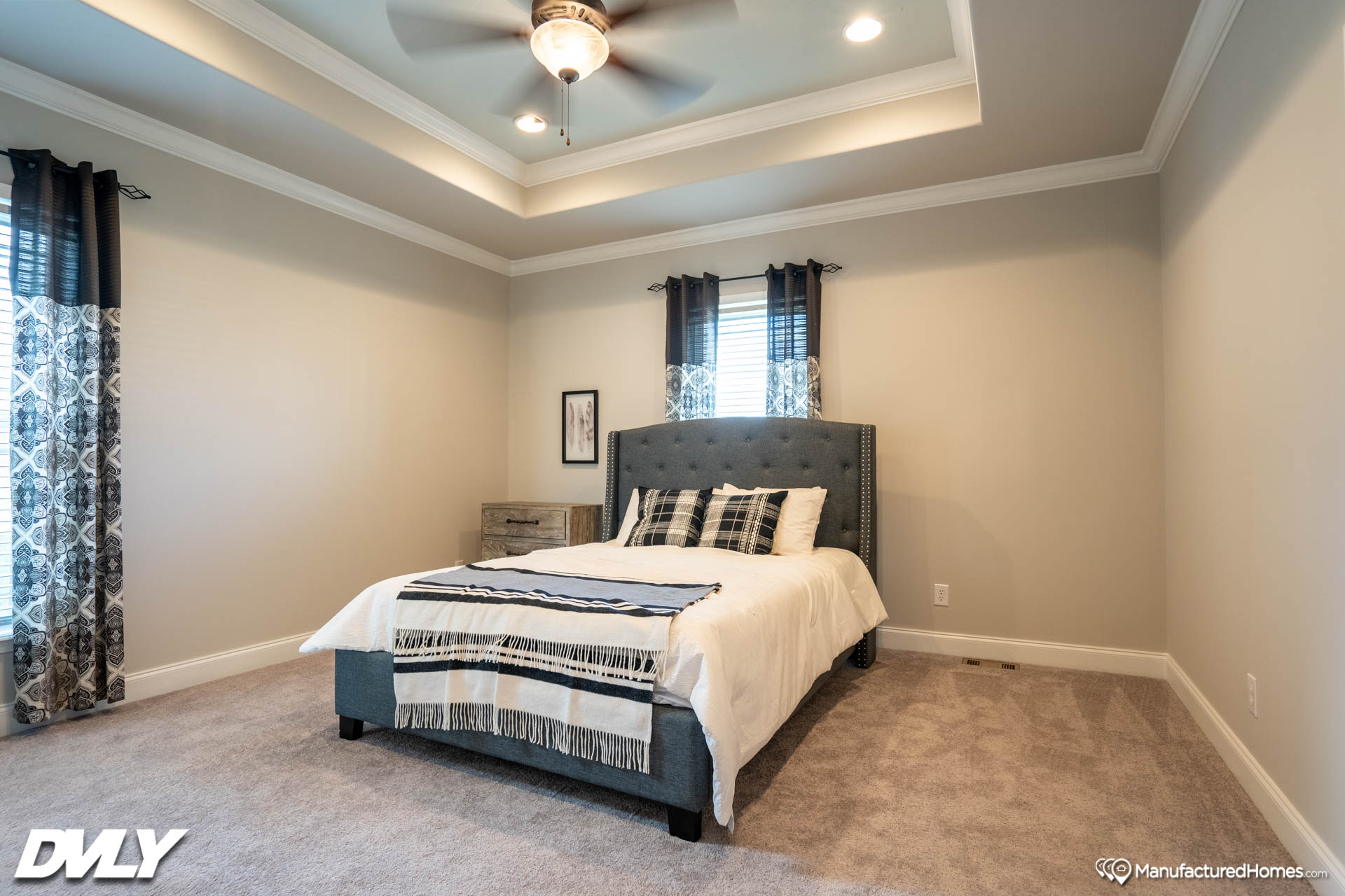 Spacious bedroom with a gray upholstered bed, white and plaid bedding. Soft lighting, patterned curtains, and a ceiling fan create a cozy, inviting atmosphere.