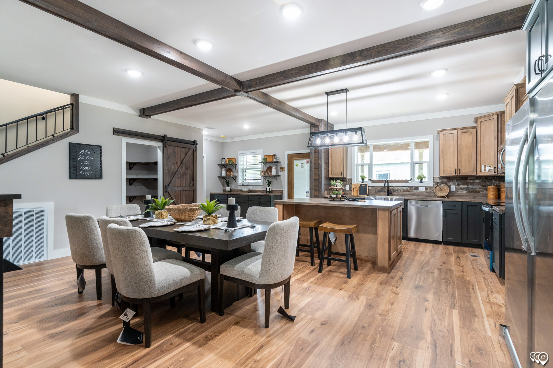 Spacious modern kitchen and dining area with light wooden flooring, exposed beams, and a rustic table set for six. Warm lighting creates a cozy ambiance.