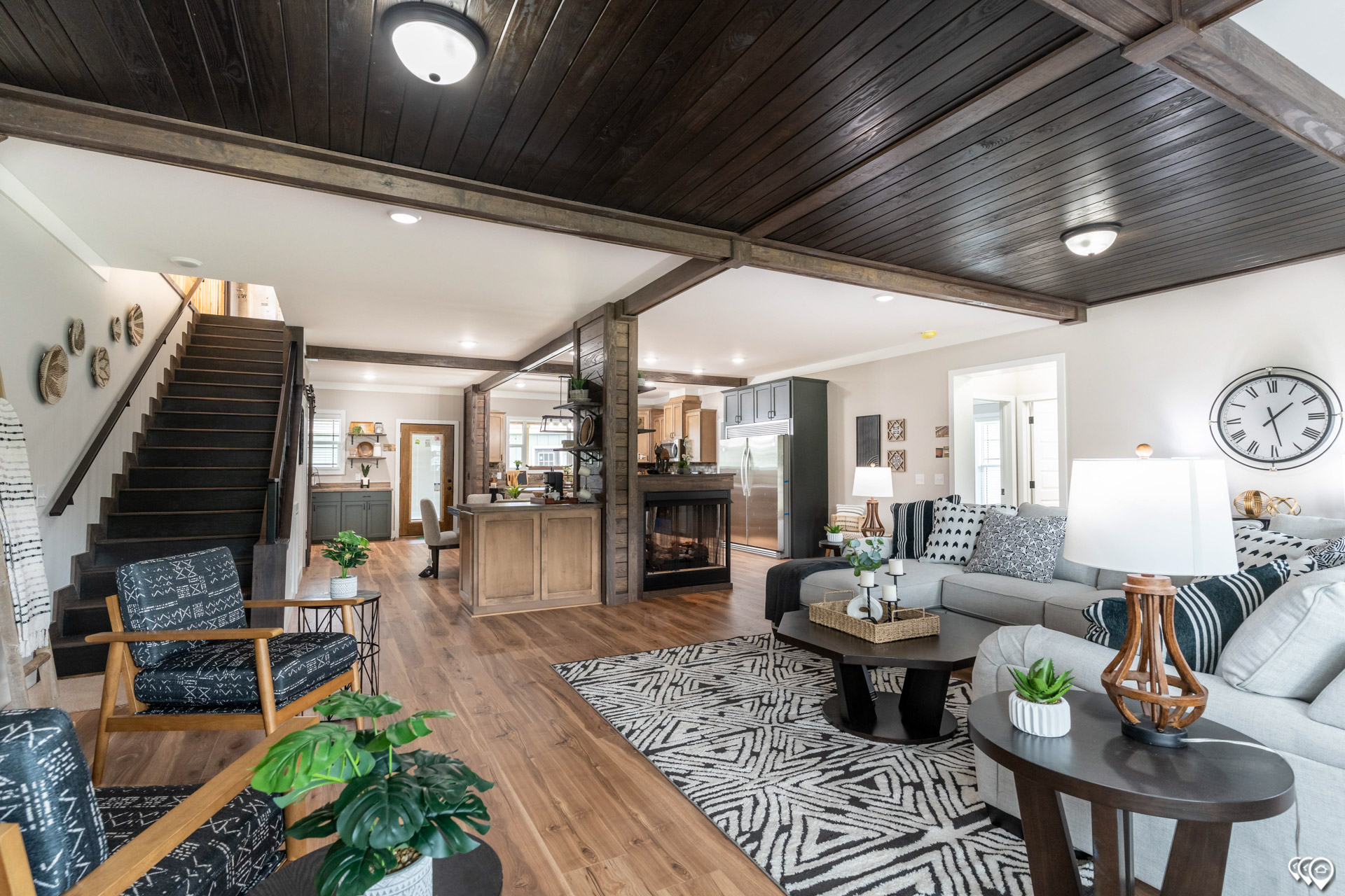 Stylish open-concept living room with wood floors, dark wood ceiling, and modern furniture. Includes a staircase, kitchen in the background, and cozy decor.