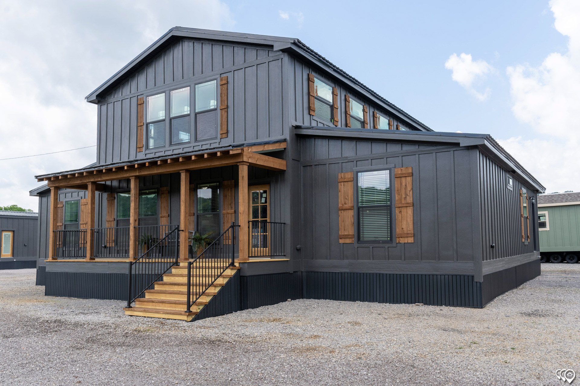 A modern, dark gray two-story house with wood accents and a covered porch. It has multiple windows with wooden shutters, set against a cloudy sky.