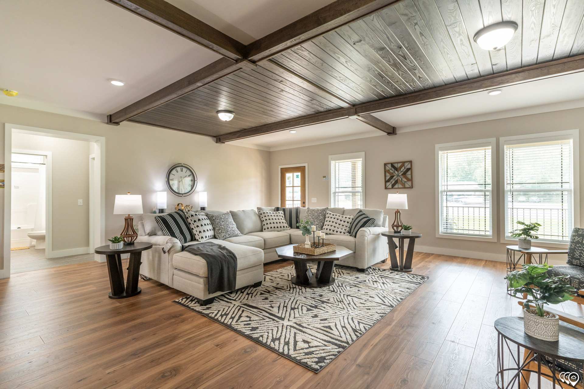 Spacious, modern living room with wood floors, beamed ceiling, and light gray sectional sofa. Decor includes patterned pillows, plants, and geometric rug.