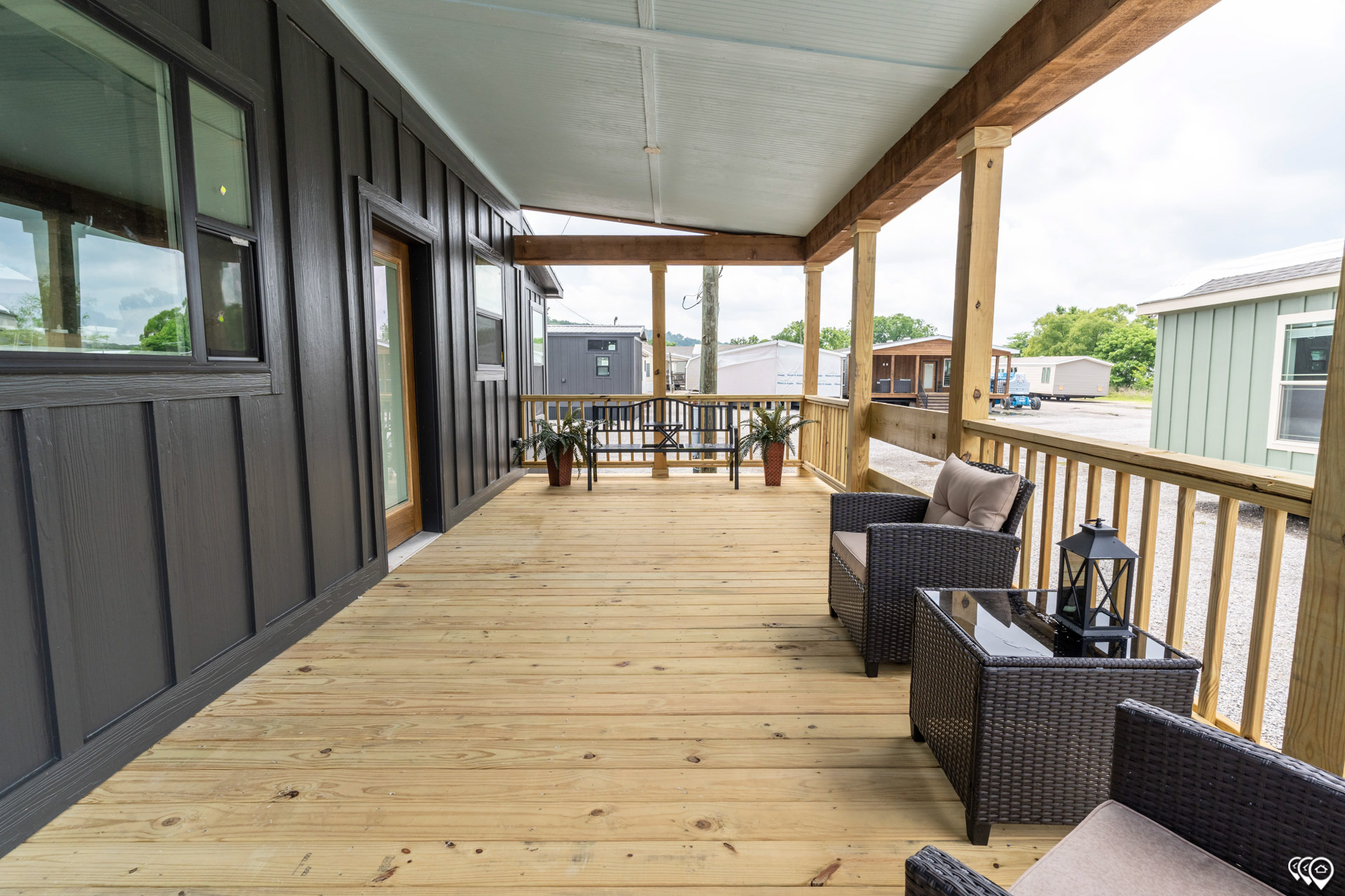 A spacious wooden porch with dark siding features wicker chairs, a glass-top table with a lantern, and potted plants, conveying a cozy, inviting atmosphere.