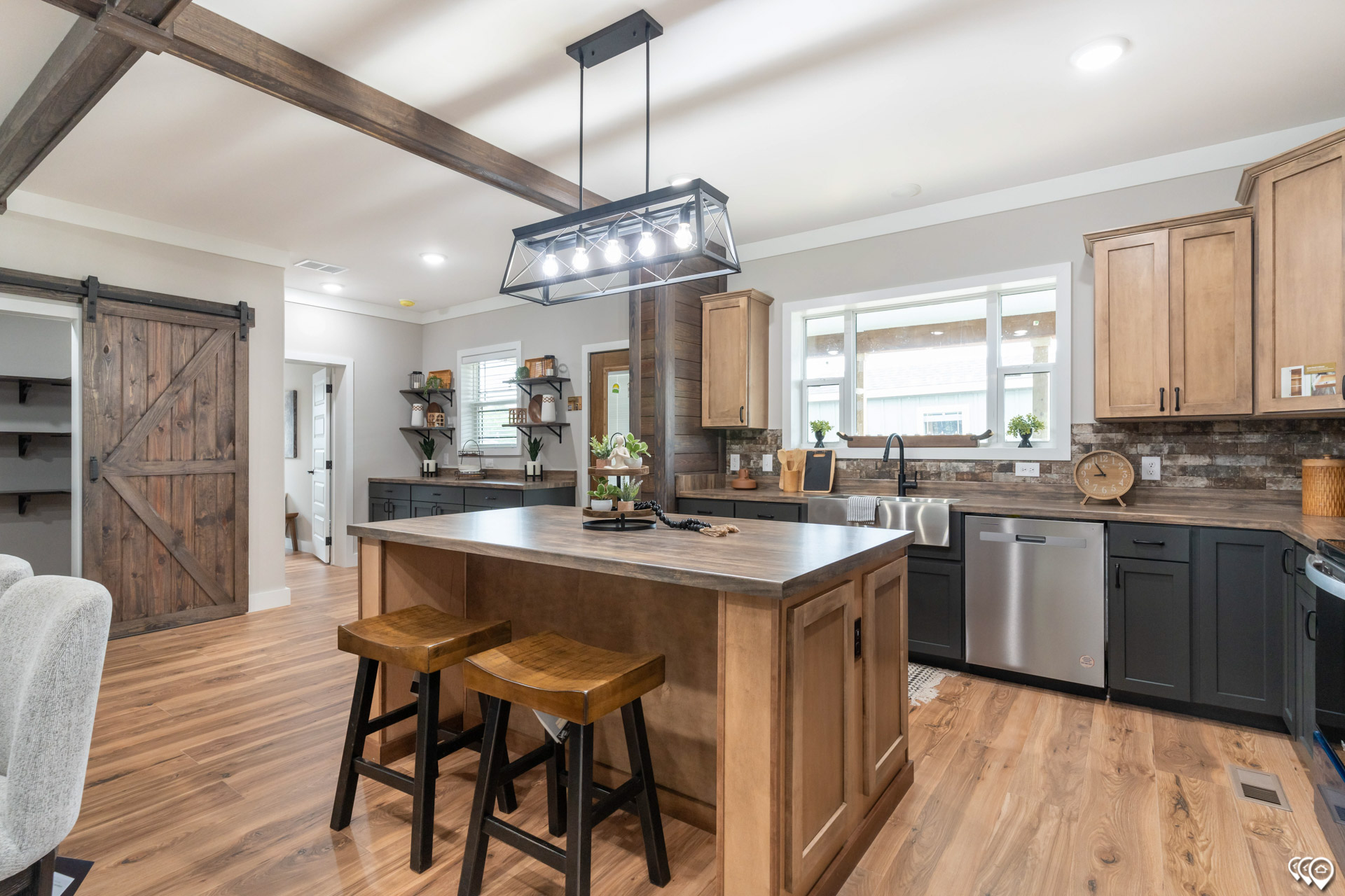 Modern farmhouse-style kitchen with wooden beams, light wooden cabinets, and a central island. Features pendant lights and rustic sliding barn door.