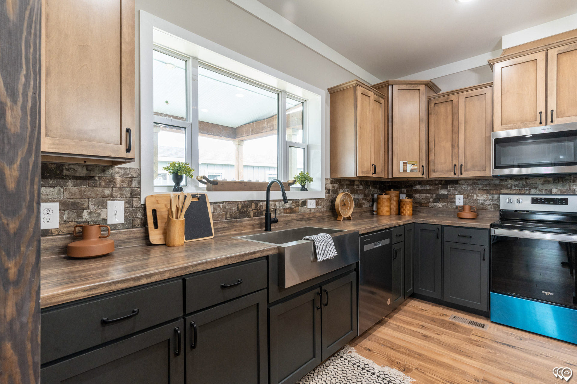 A modern kitchen with wooden cabinets, a farmhouse sink, and a black countertop. Natural light streams through a large window, creating a warm ambiance.
