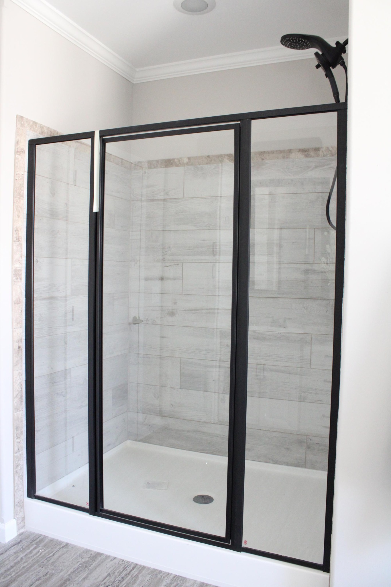 Modern shower with clear glass doors framed in black. The interior features light gray, textured tiles. A sleek showerhead is visible above.