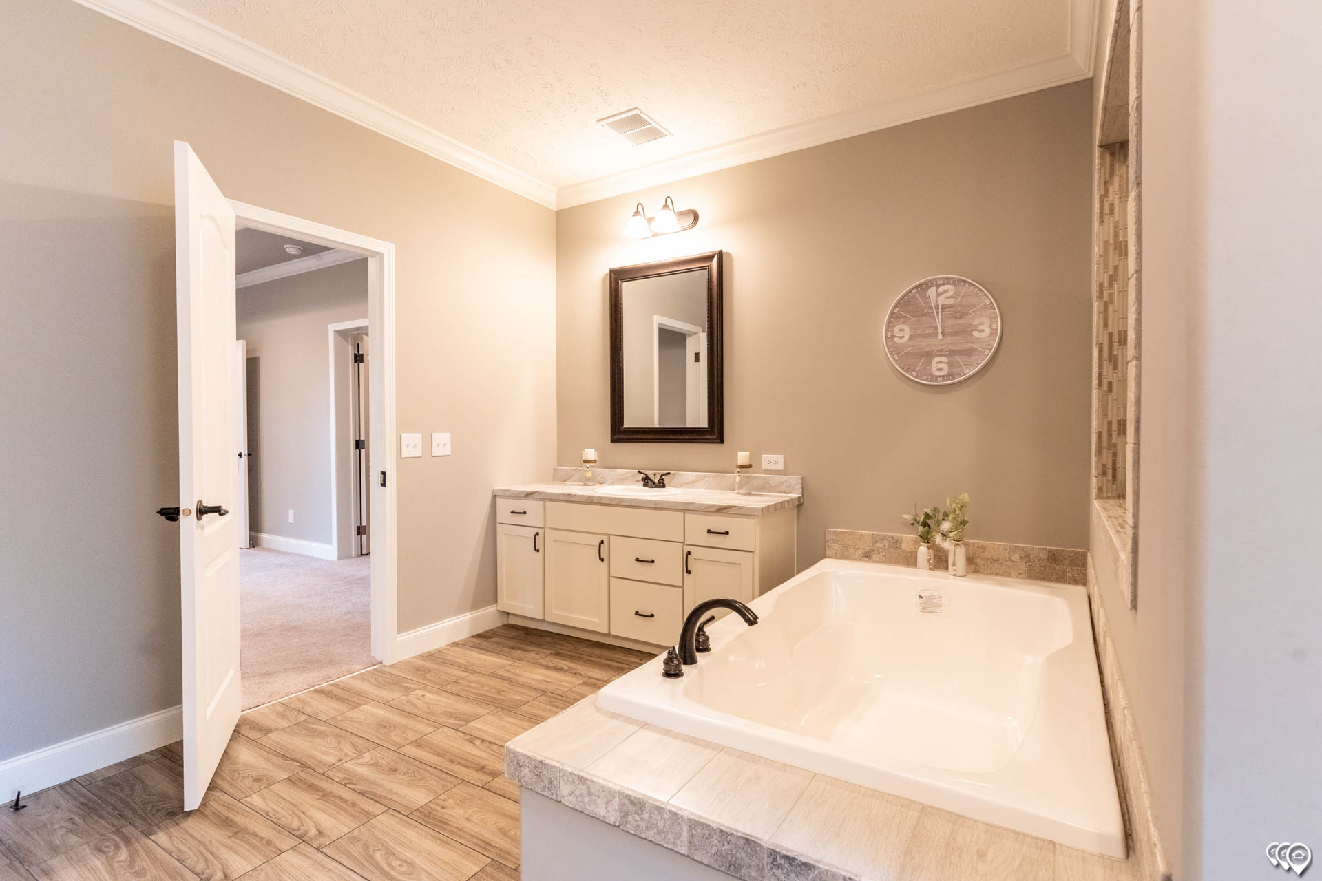 Spacious bathroom with beige walls, featuring a large white bathtub with black fixtures, a vanity with a mirror, wooden floor, and open door.