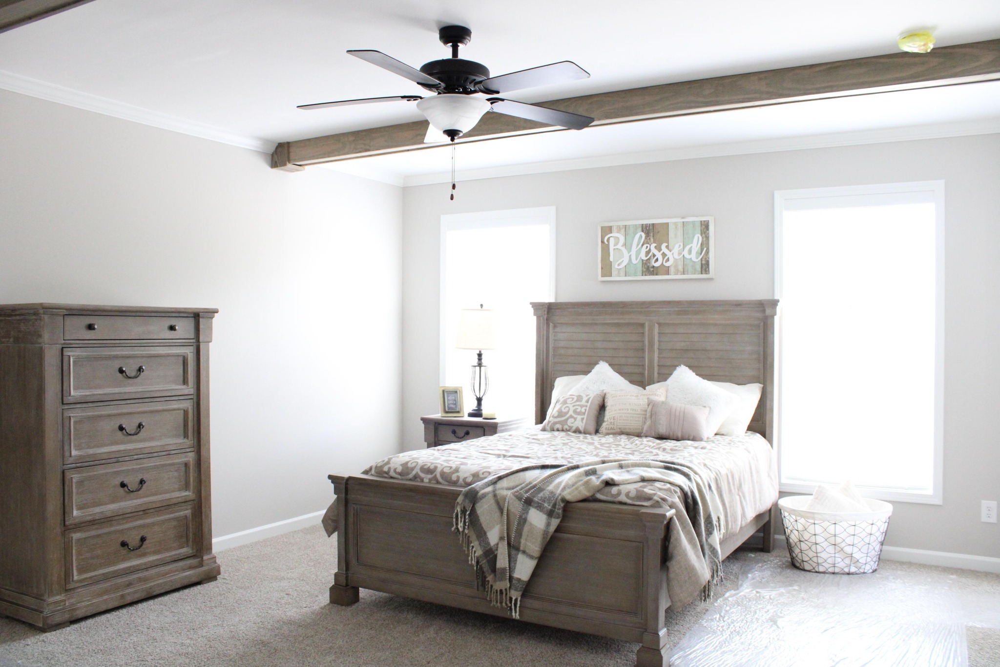 A cozy bedroom with neutral tones features a wooden bed, plaid blanket, and pillows. A dresser and basket are on a soft carpet. A "Blessed" sign and a ceiling fan add charm.