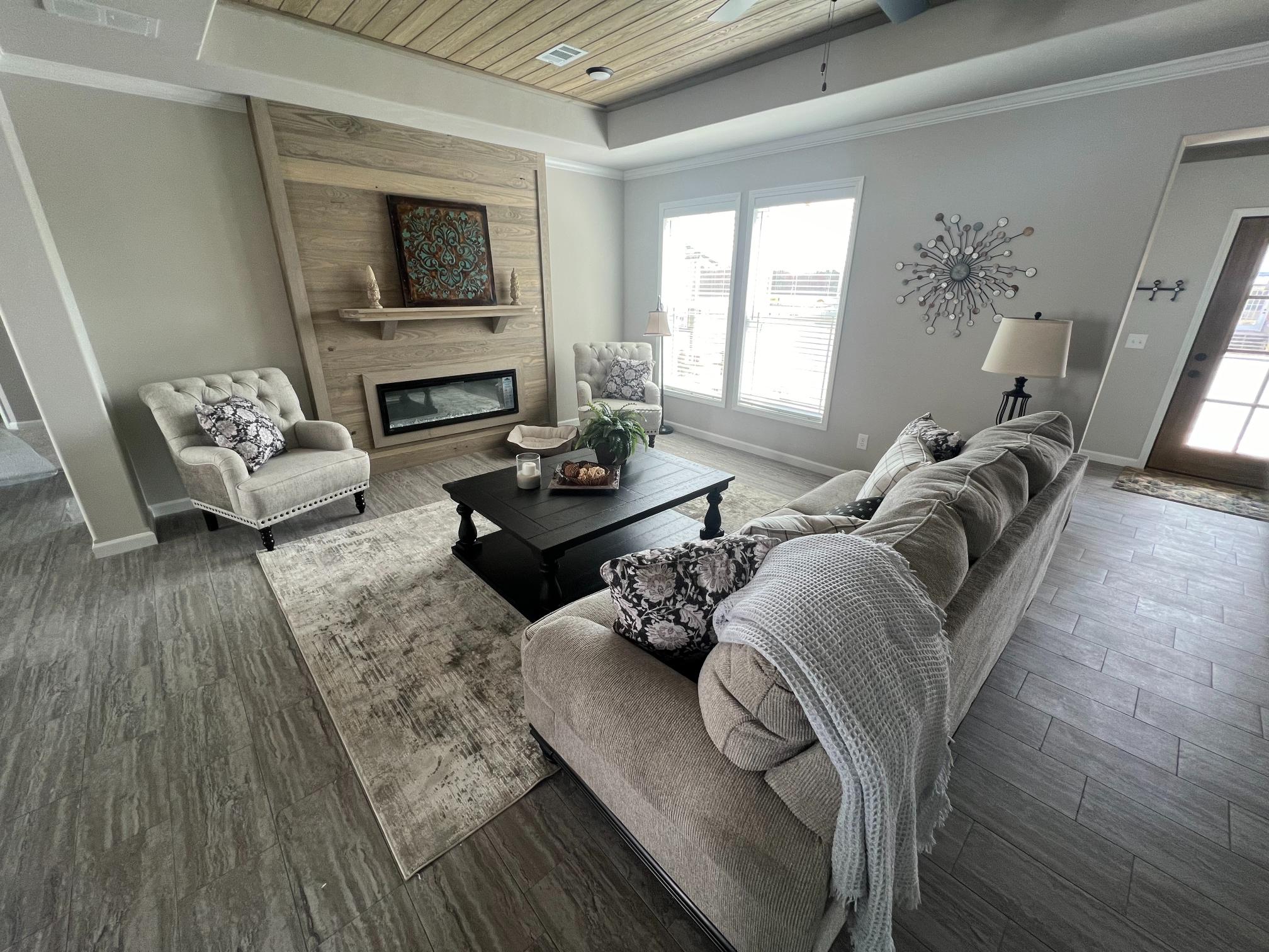A modern living room with a cozy beige couch adorned with patterned cushions and a white throw. Two tufted chairs face a black coffee table holding candles and a plant. A rustic fireplace with wood paneling and decorative art is central, flanked by windows letting in natural light. A decorative wall clock and lamp enhance the inviting, serene atmosphere.
