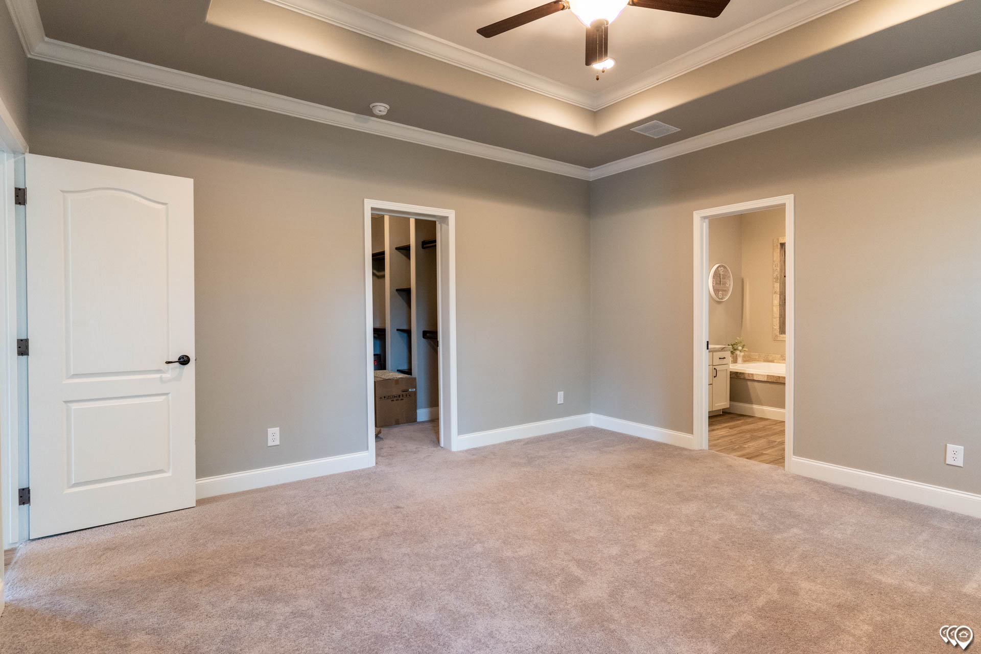Spacious beige carpeted room with tray ceiling and ceiling fan. Two doorways lead to a closet and bathroom, enhancing the sense of openness and functionality.