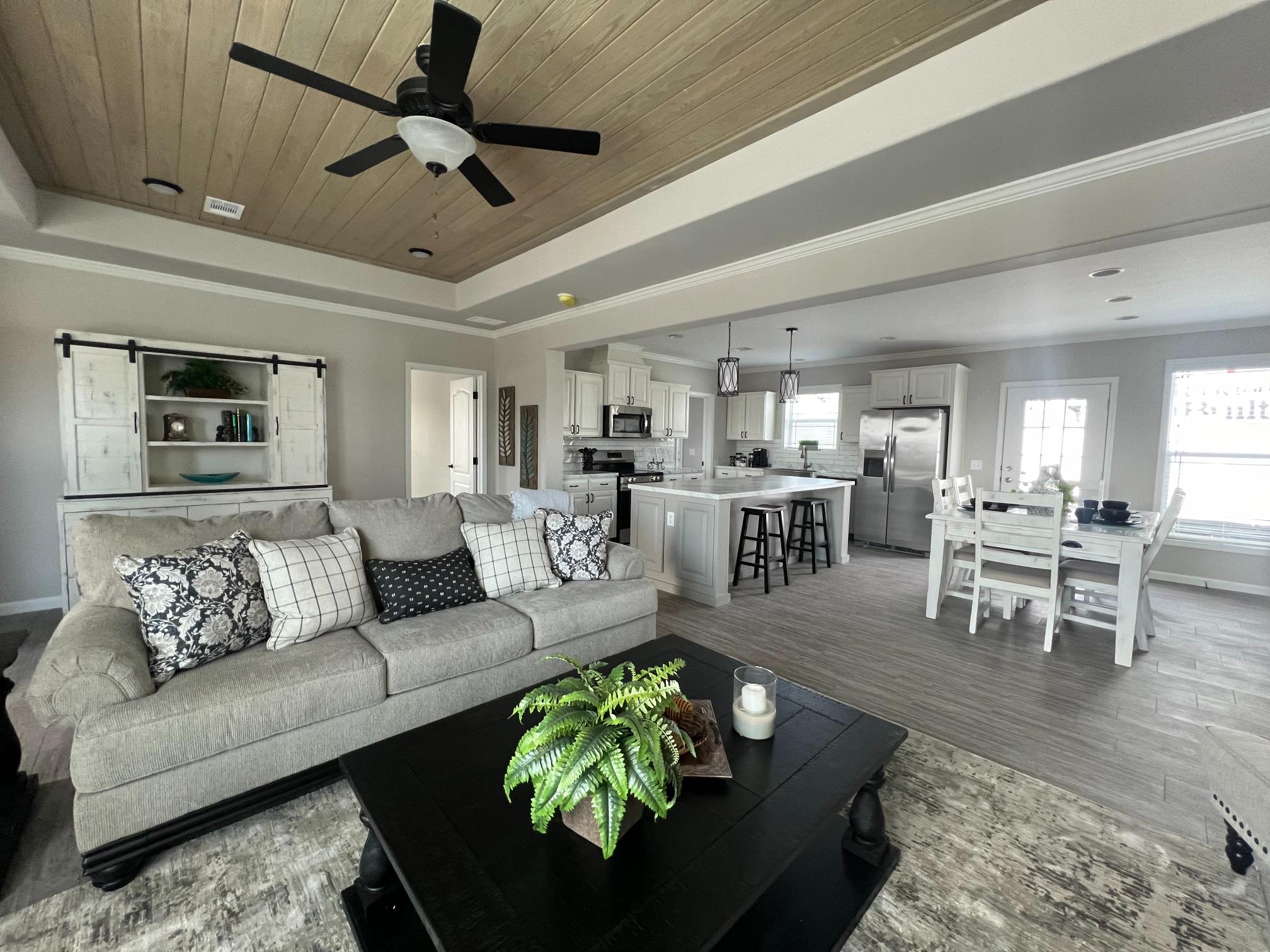 Modern living room open to kitchen, featuring a beige sofa with patterned pillows, black coffee table with plant, dining area, and ceiling fan. Bright and inviting.