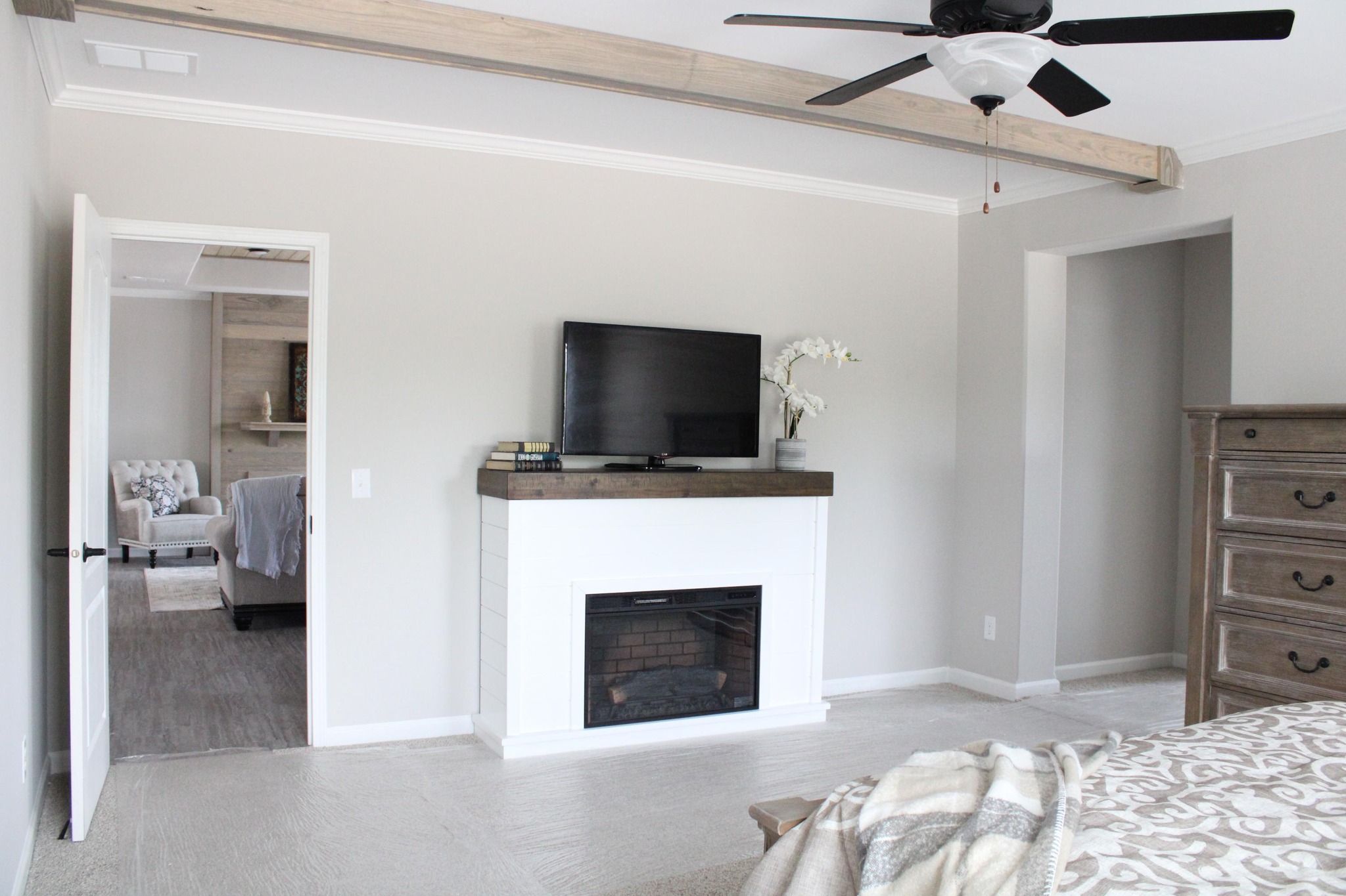 Bedroom with neutral tones, featuring a wall-mounted TV above a white fireplace. A ceiling fan adds comfort. An open door reveals a cozy seating area.