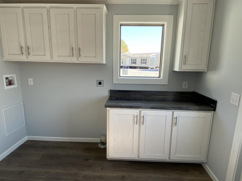 A small kitchen space with white cabinets and a dark countertop beneath a window. Natural light highlights the light gray walls and dark wood floor.