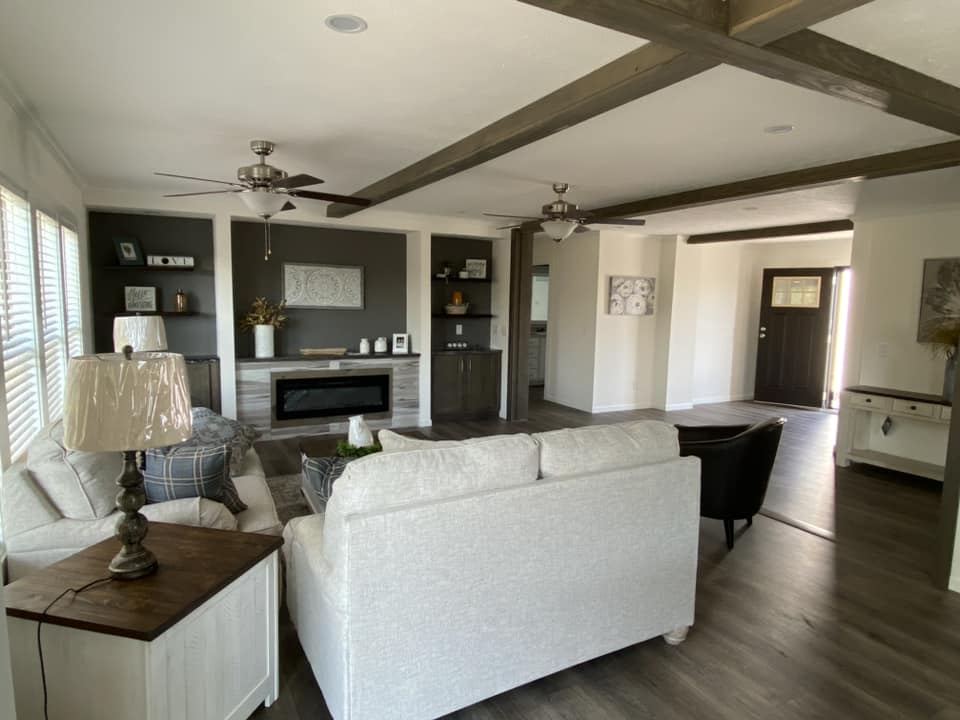 A modern living room with light gray sofas, a wooden coffee table, and a fireplace. Dark wood beams on the ceiling, fans, and a large window add warmth.