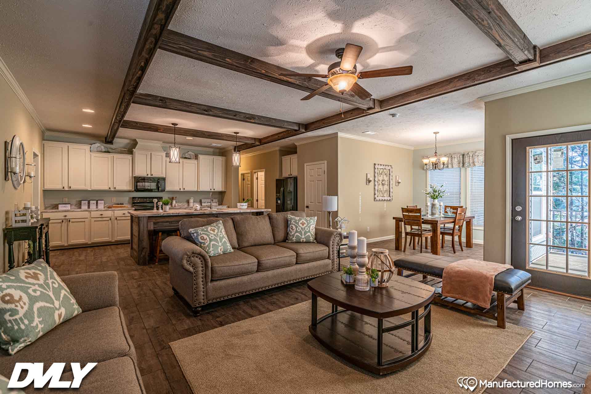 Spacious living room with beige sofas, wooden beams, and ceiling fan. Open layout leads to a kitchen and dining area. Warm, cozy atmosphere.