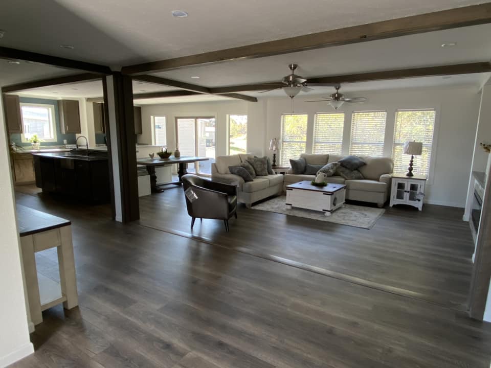 Spacious living area with dark wood flooring, beige sofas, and large windows. Open kitchen with an island to the left and modern decor. Bright and inviting.
