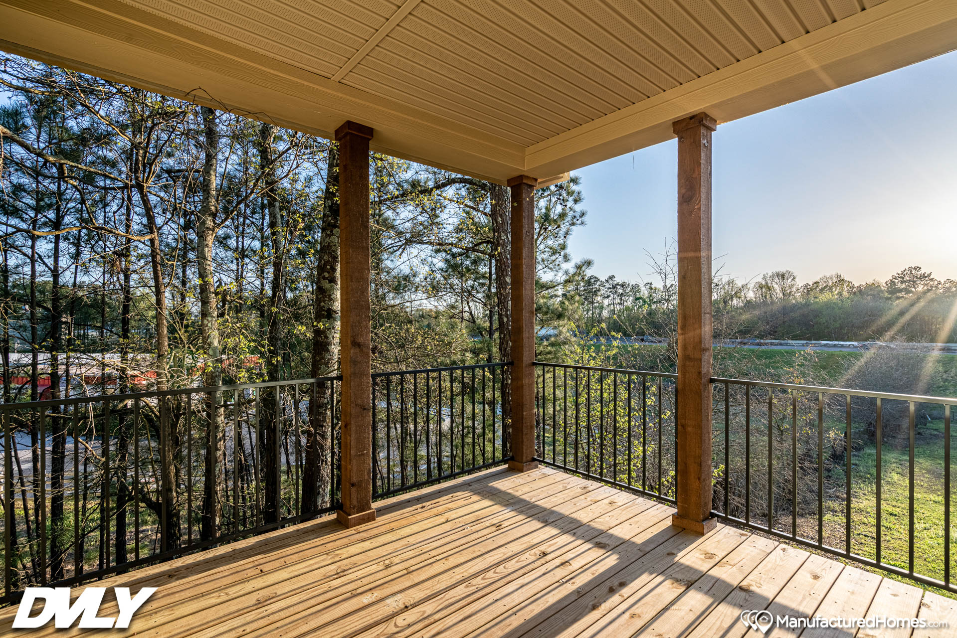 A serene wooden deck with railings overlooks a lush, green forest. Sunlight filters through trees, casting long shadows, creating a tranquil, cozy atmosphere.