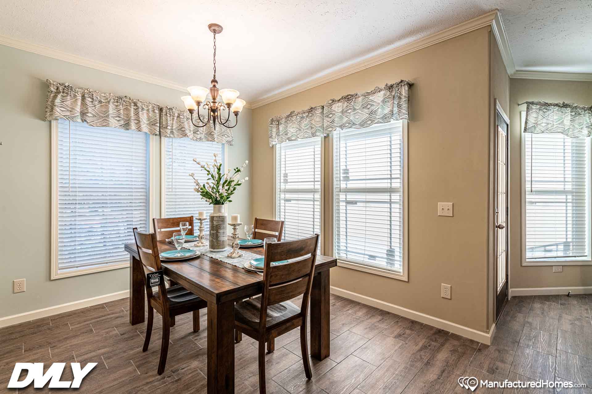 A cozy dining room with wooden table set for four, turquoise accents, and floral centerpiece. Light streams through large, curtained windows, creating a warm ambiance.
