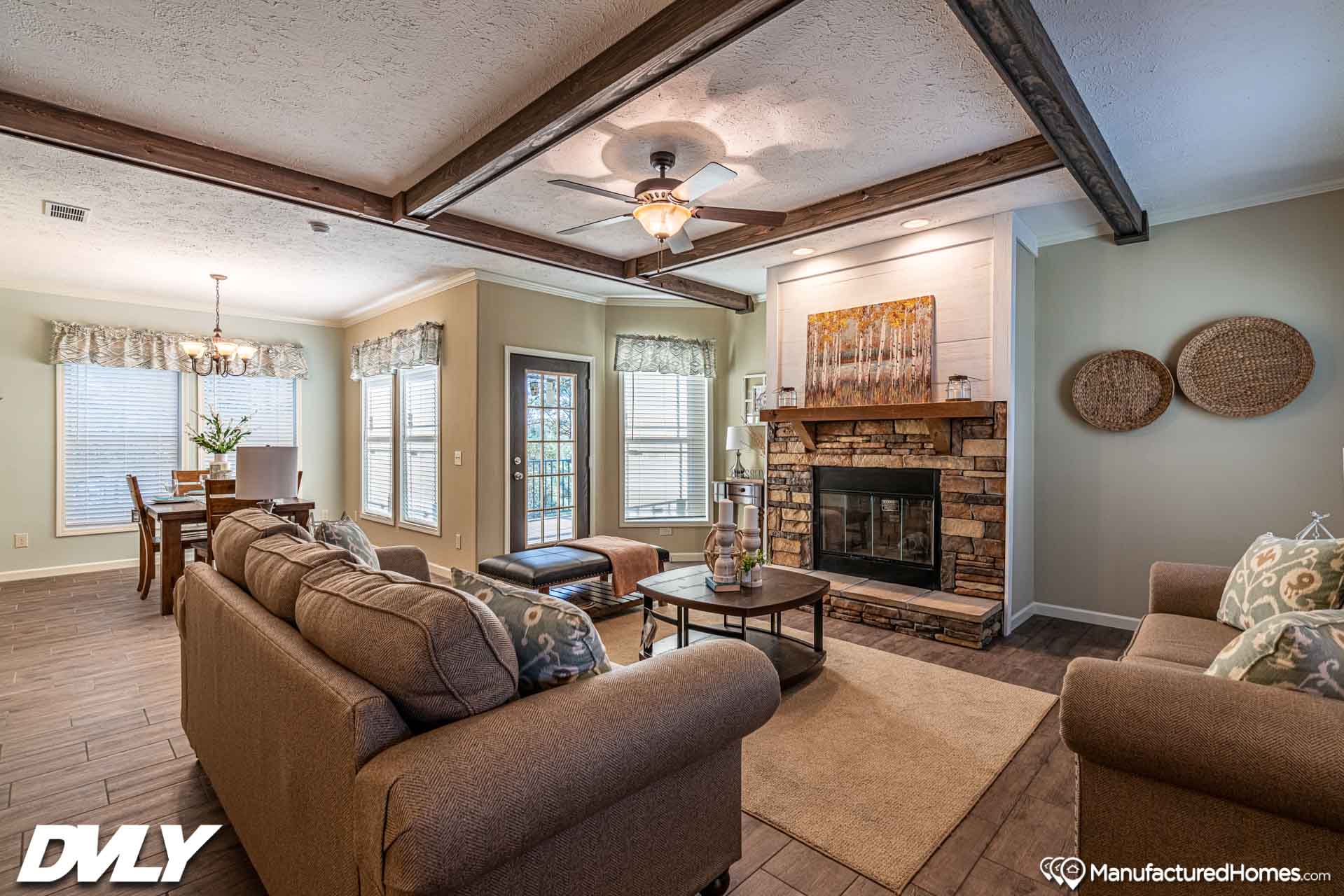 Cozy living room with a brown sofa set, brick fireplace, and beamed ceiling. A dining area is visible, creating a warm, inviting atmosphere.