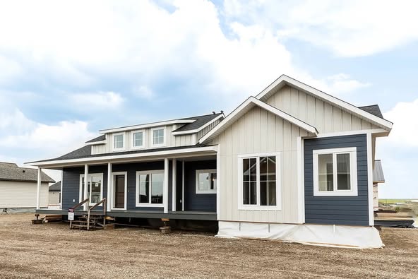 Modern farmhouse with blue and white siding and a steep roof. It features large windows and a spacious porch, set under a partly cloudy sky.