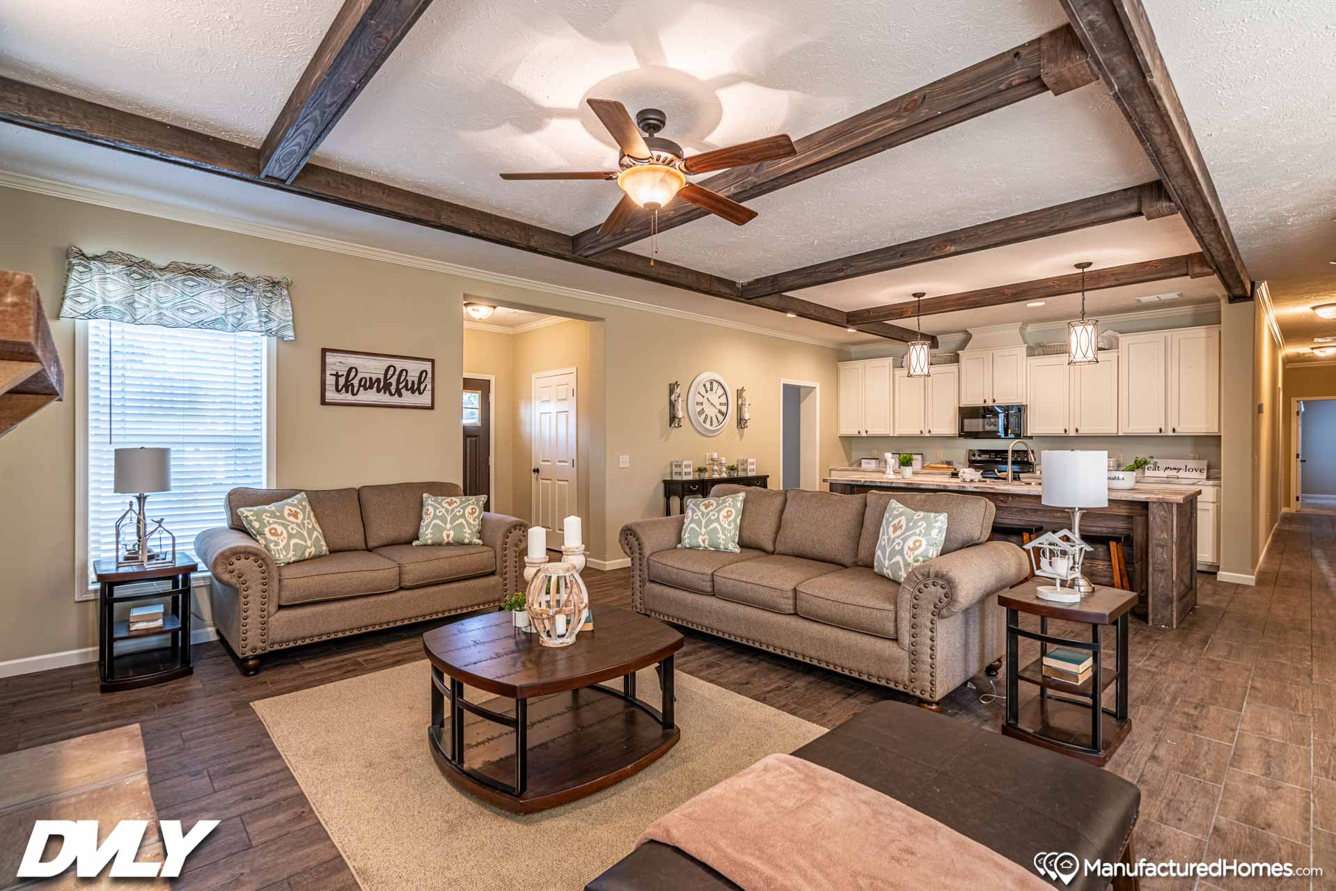 Spacious living room with brown sofas, a coffee table, and ceiling fan. Open kitchen in background with white cabinets. Warm, cozy atmosphere.