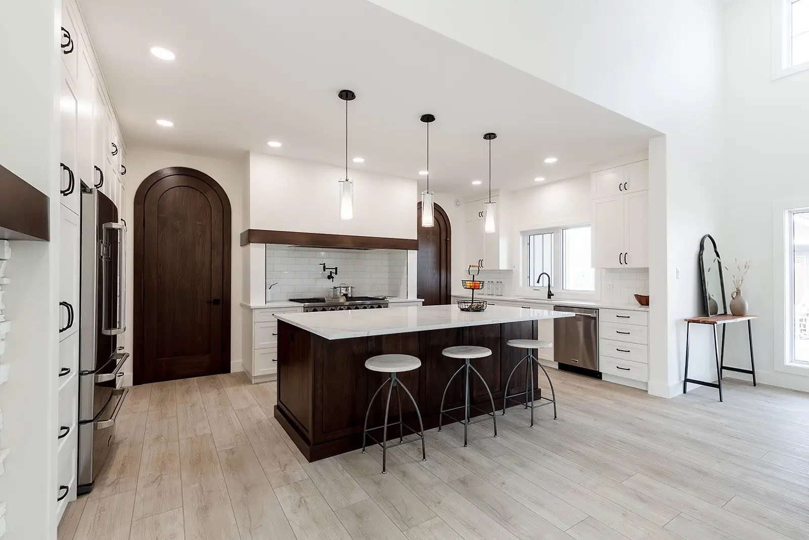 Modern kitchen with white walls, wooden floors, and dark cabinetry. Features an island with stools, pendant lighting, and stainless steel appliances.