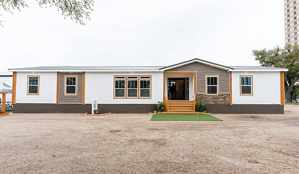 Single-story, modern manufactured home with a white facade, wooden accents, and multiple windows. A welcoming porch with steps and greenery is front center.
