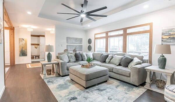 Modern living room with a large gray sectional sofa, green cushions, and a central ottoman. Natural light streams through wood-framed windows, creating a cozy ambiance.