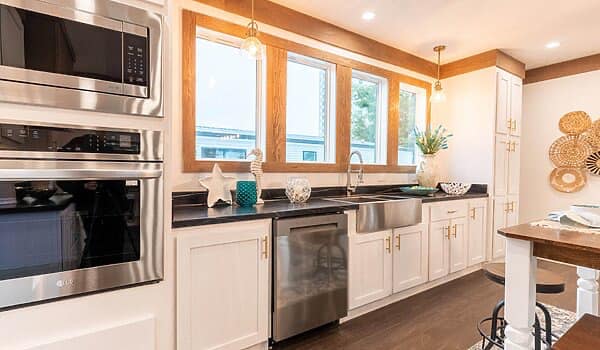 Bright kitchen with white cabinets, stainless steel appliances, and a farmhouse sink. Three windows, decorative items, and pendant lights enhance a cozy, inviting atmosphere.