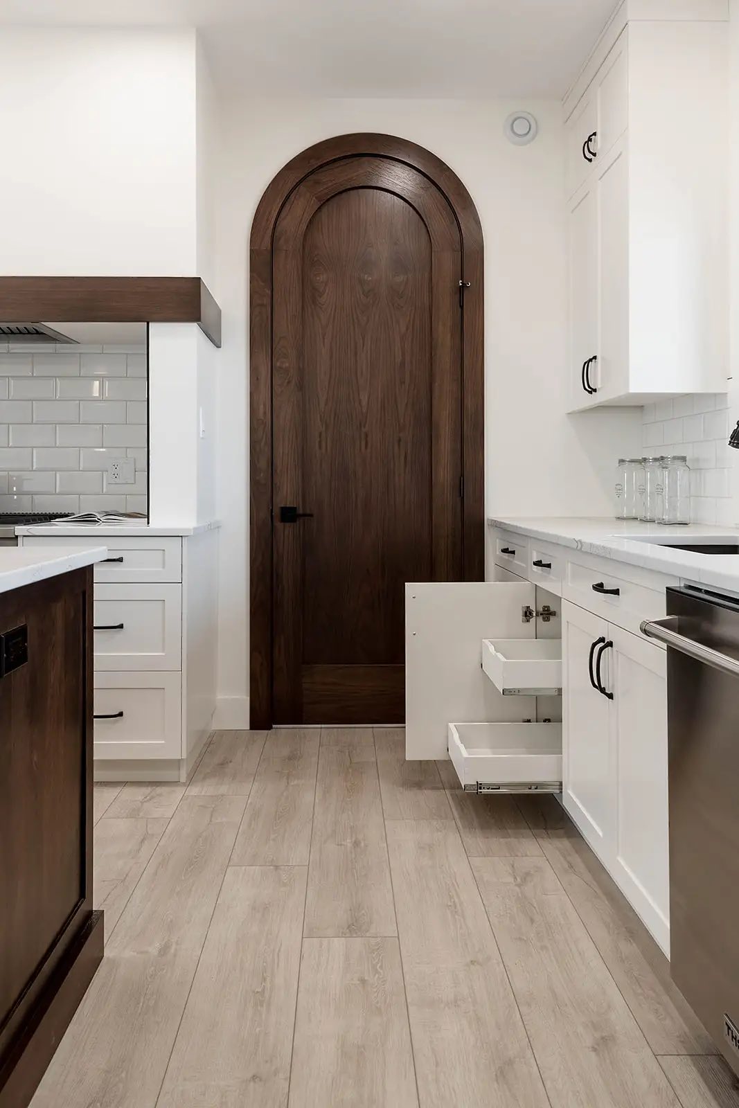 Modern kitchen with light wood flooring, a tall arched wooden door, white cabinets, black handles, open drawers, and subway tile backsplash.