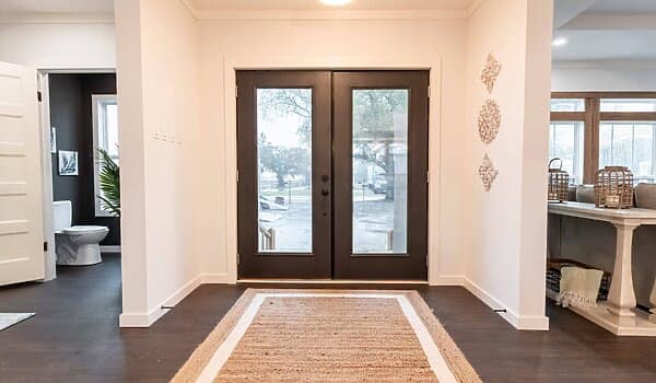 Modern entryway with dark double doors, large windows, and a woven rug on wooden floors. A bathroom is visible on the left, and decor accents on the wall.