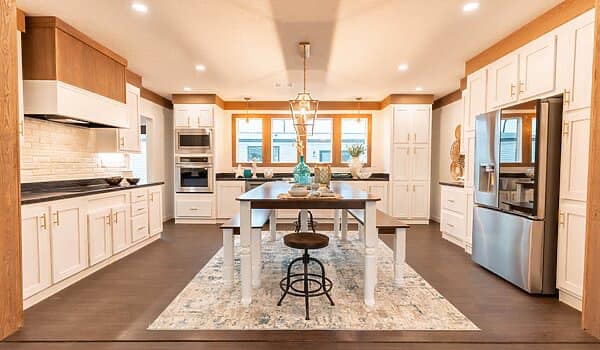 A modern kitchen with sleek white cabinets, stainless steel appliances, and a central wooden table on a patterned rug. Warm lighting and decor create a cozy atmosphere.