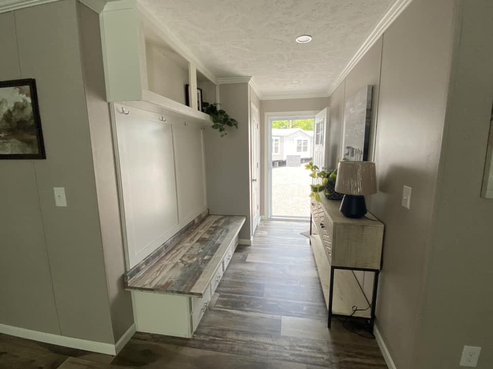A cozy hallway with light gray walls and laminate wood flooring. It features a wooden bench with hooks above, a console table with plants, and a lamp. Bright and welcoming ambiance.