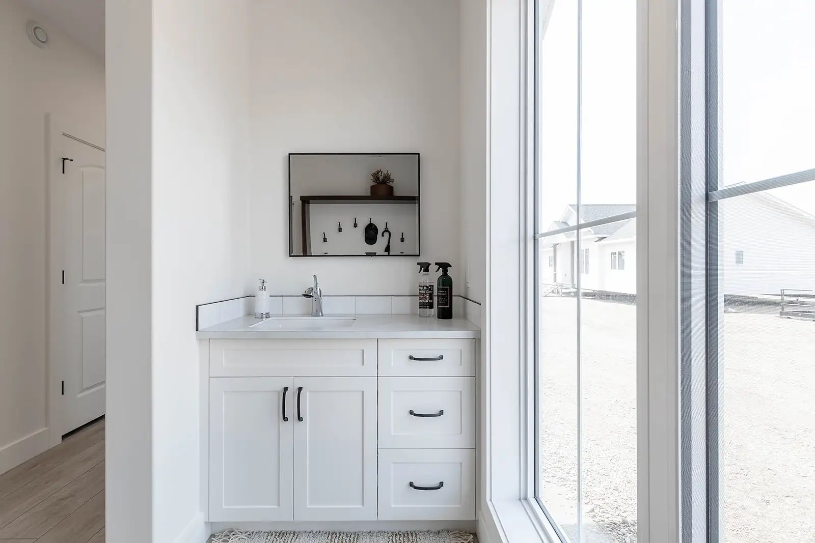 A minimalist white bathroom with a vanity sink, black handles, and a wall-mounted mirror. Natural light streams through a large window, creating a serene atmosphere.