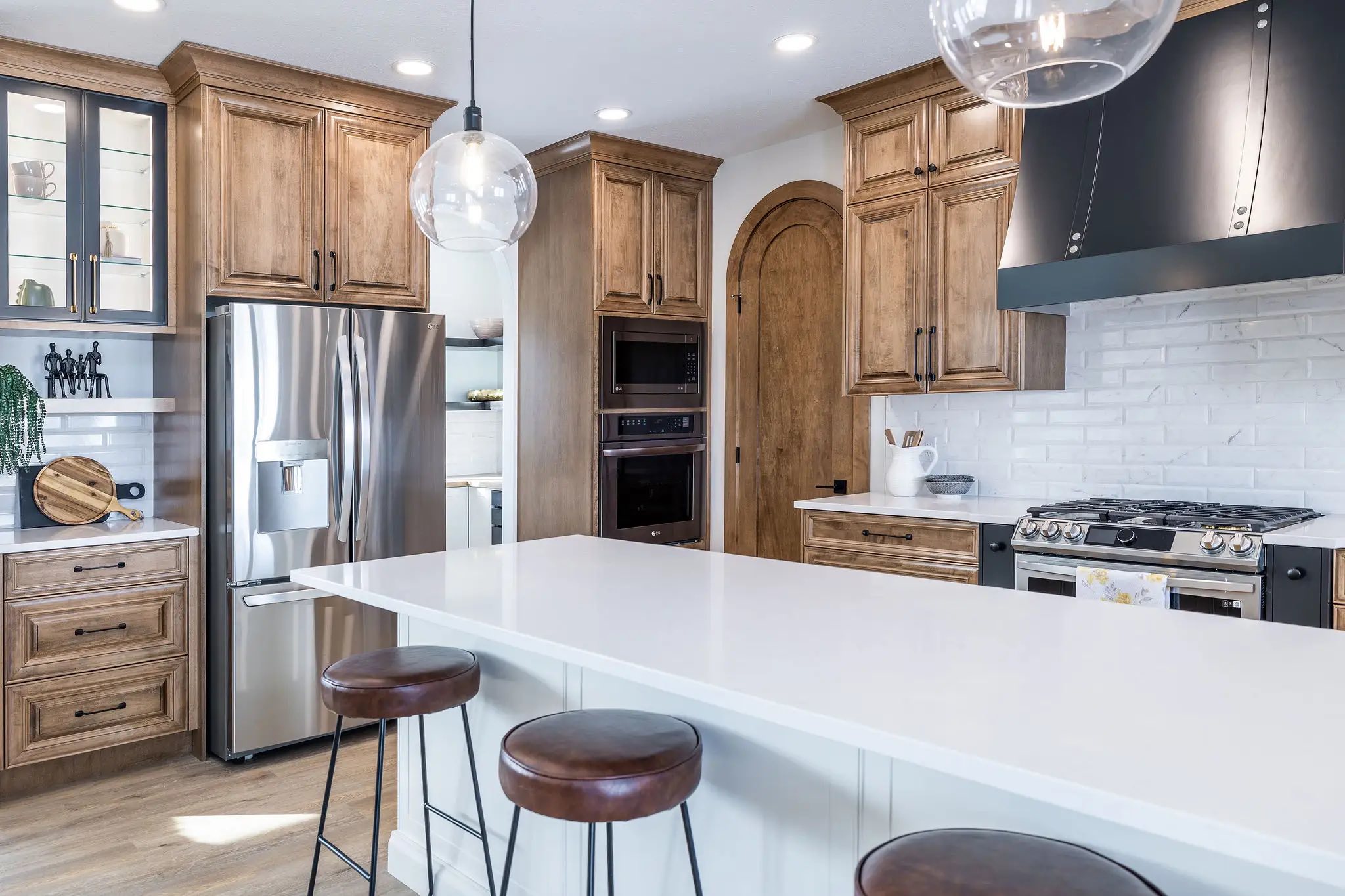 Modern kitchen with wooden cabinets and stainless steel appliances. A large white island with brown stools sits under stylish pendant lights. Bright and elegant atmosphere.