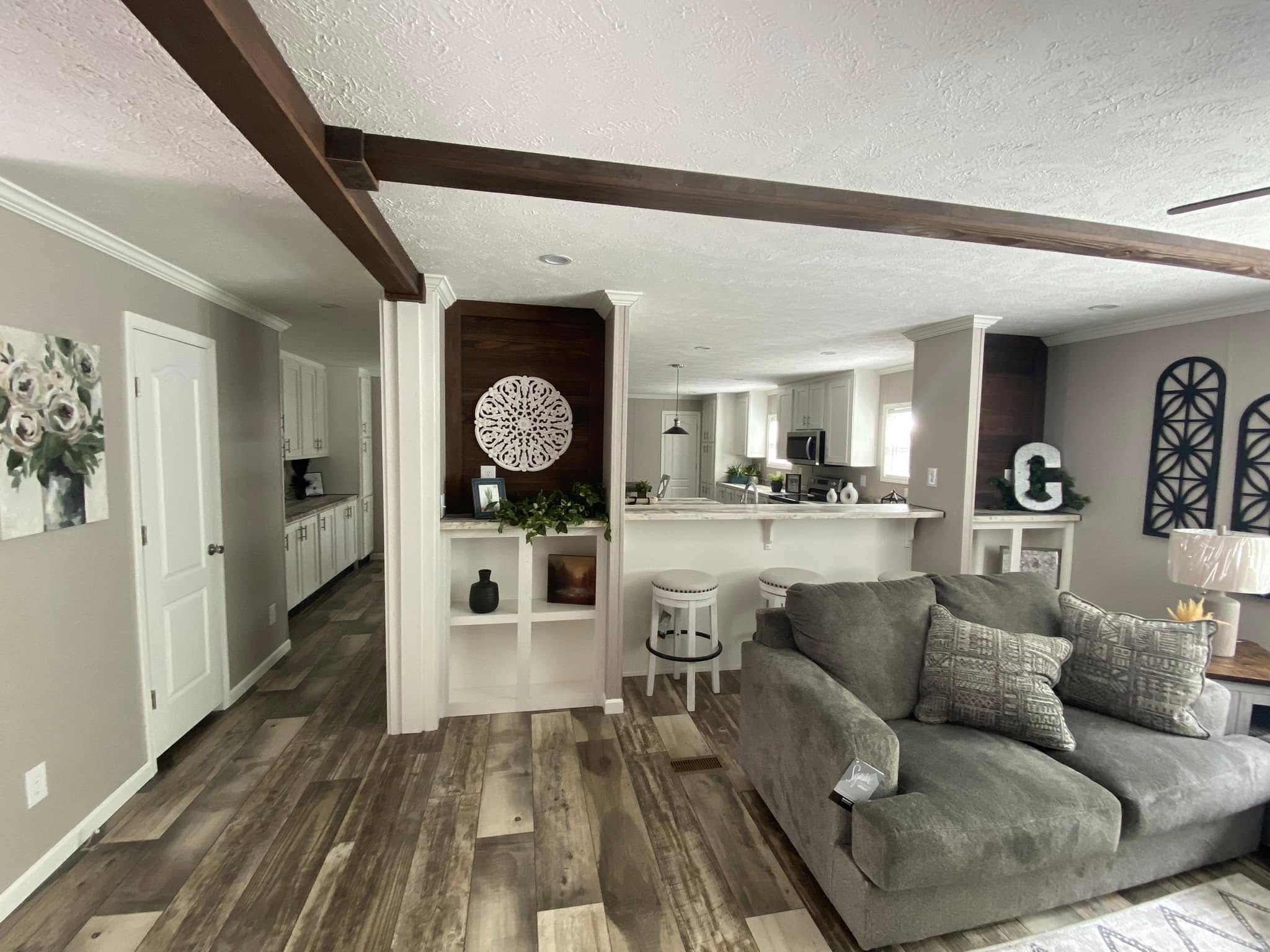 Spacious living area with a gray couch, wood floors, and white walls. Open kitchen features bar stools and pendant lighting, creating a cozy, inviting ambiance.