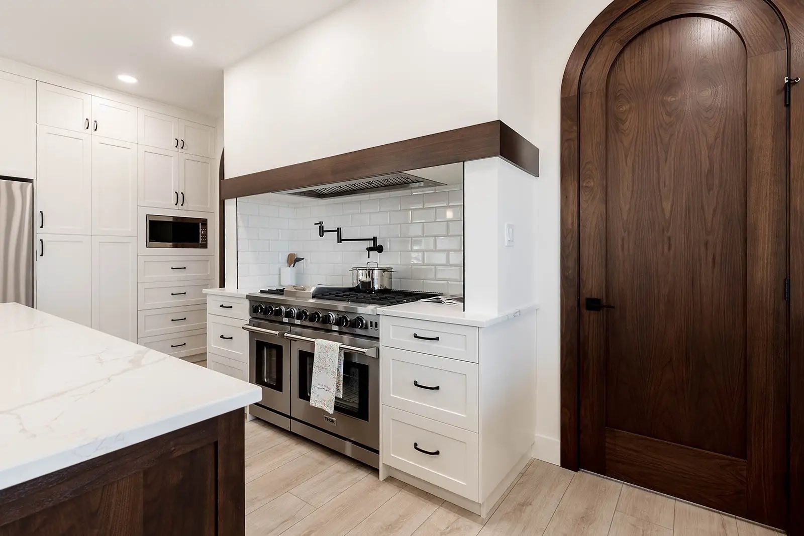 Sleek kitchen with white cabinetry, stainless steel oven, and subway tile backsplash. Features a large arched wooden door and modern appliances.