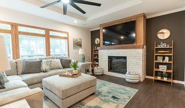 Cozy living room with a large sectional sofa, patterned rug, and a modern ceiling fan. A flat-screen TV is mounted above a white brick fireplace.