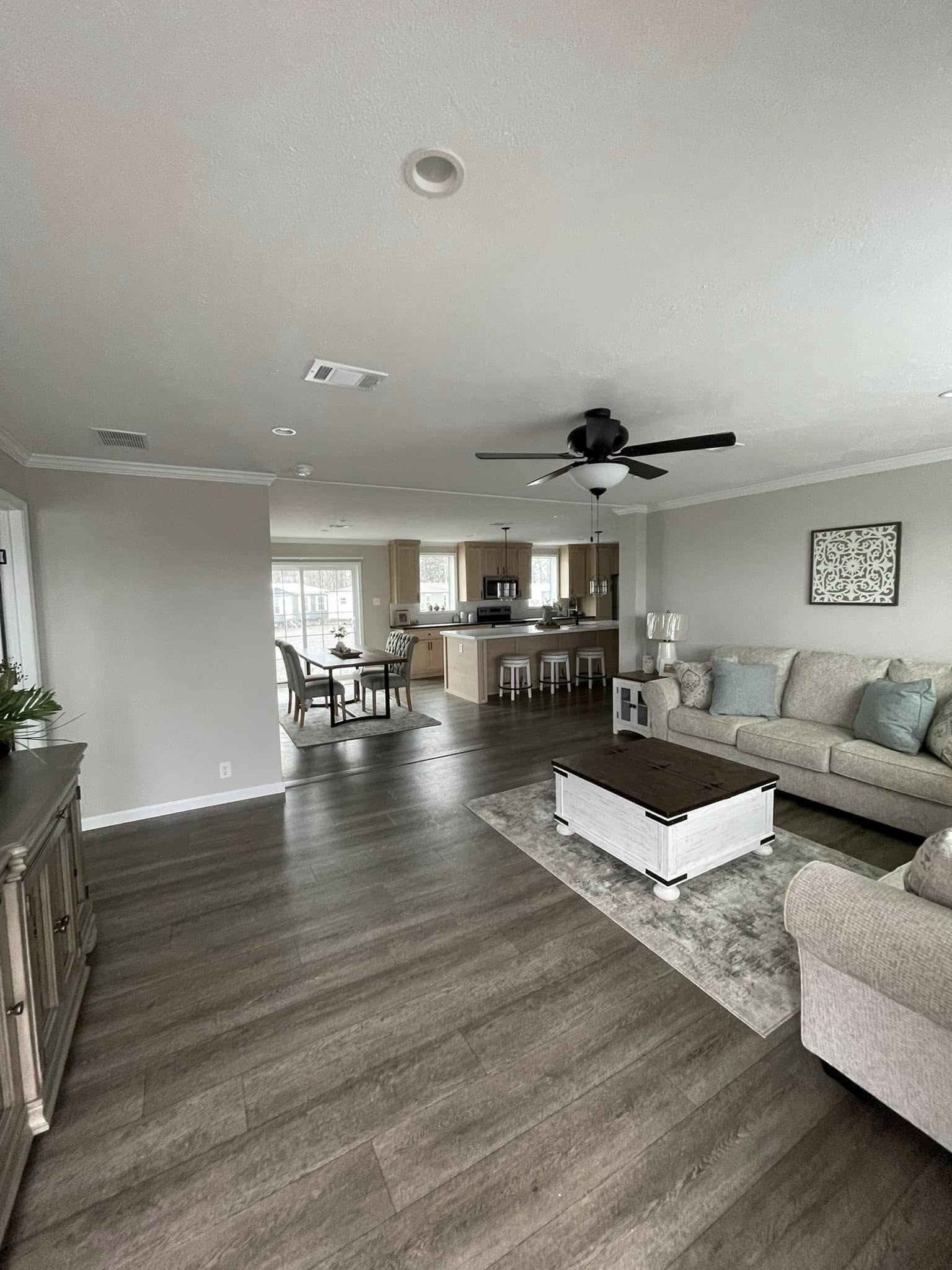 Spacious living room with gray walls and wood flooring, featuring a cozy beige sofa with blue cushions, a white coffee table, and a ceiling fan. Opens to a modern kitchen with an island and chairs, creating an inviting atmosphere.