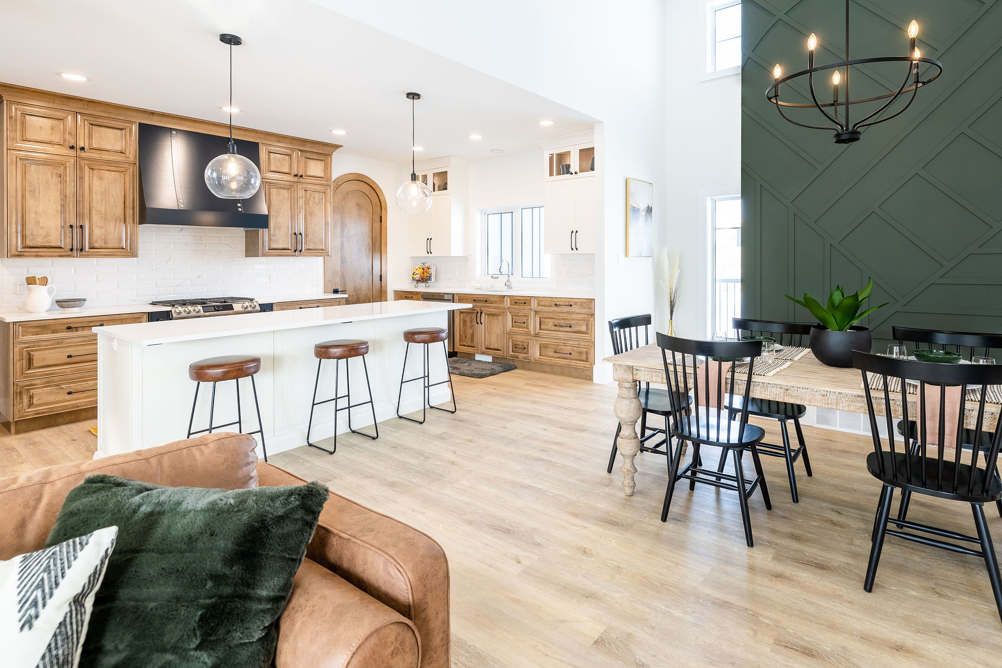 Modern kitchen and dining area featuring wood cabinets, a white island with four stools, black chairs around a table, and a green accent wall. Airy and inviting atmosphere.