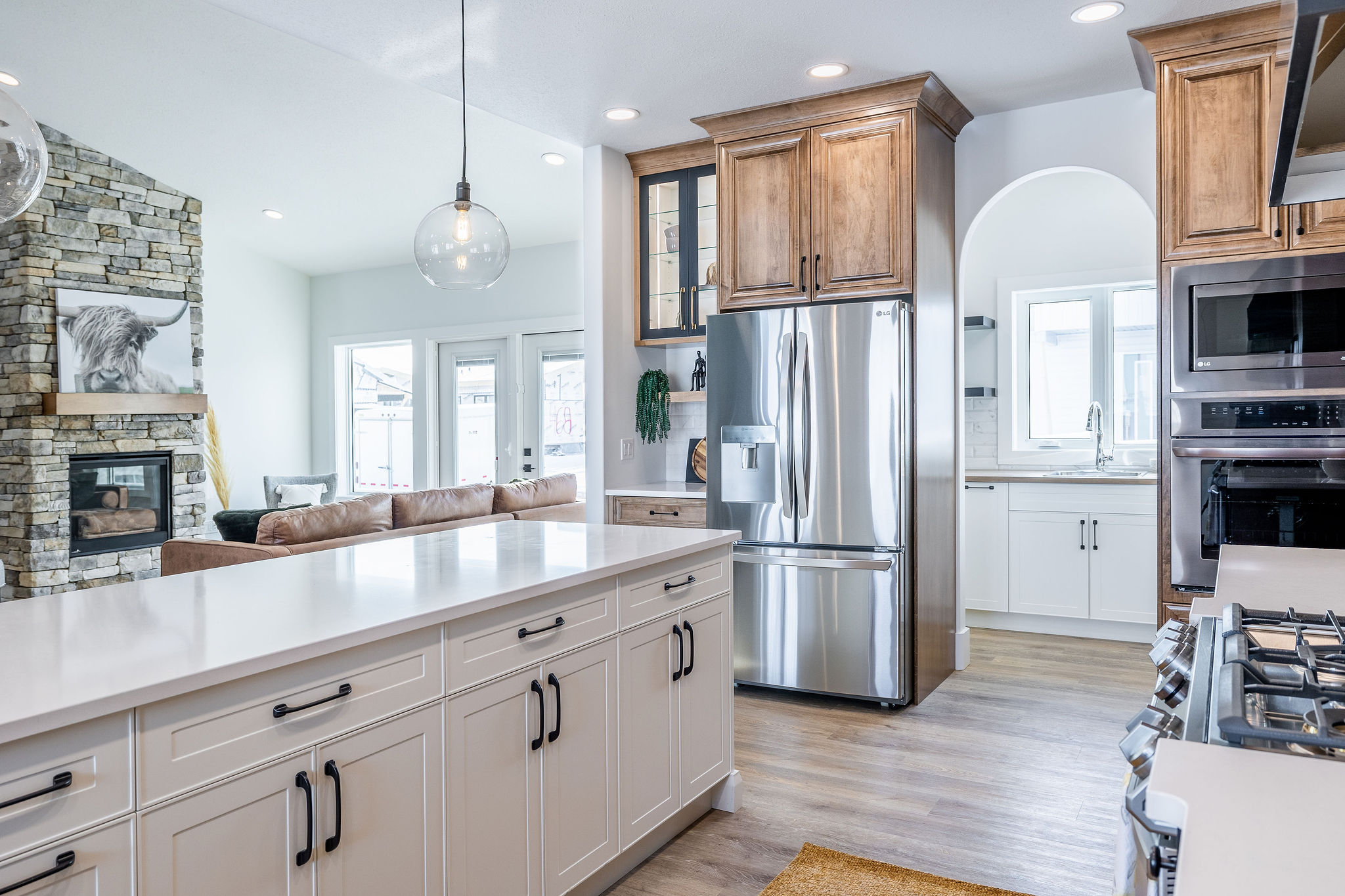 Modern kitchen with white cabinetry, stainless steel appliances, and wooden accents. Stone fireplace, cozy living room visible in the background. Bright and airy.